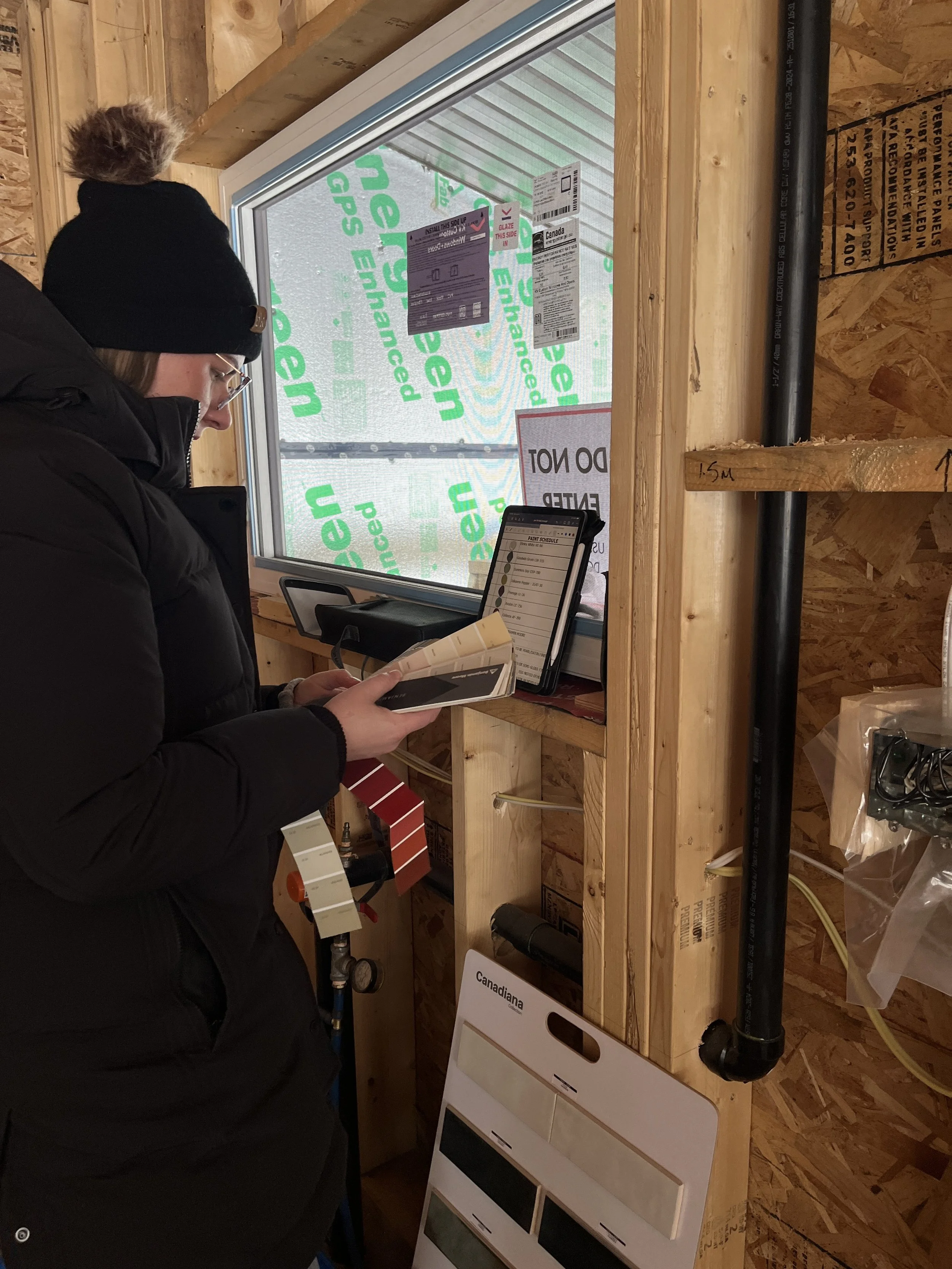 A woman in a black coat and beanie looks at a color sample book inside a partially constructed building. The interior features wooden framing with insulation and a window, through which construction labels and safety signs are visible.