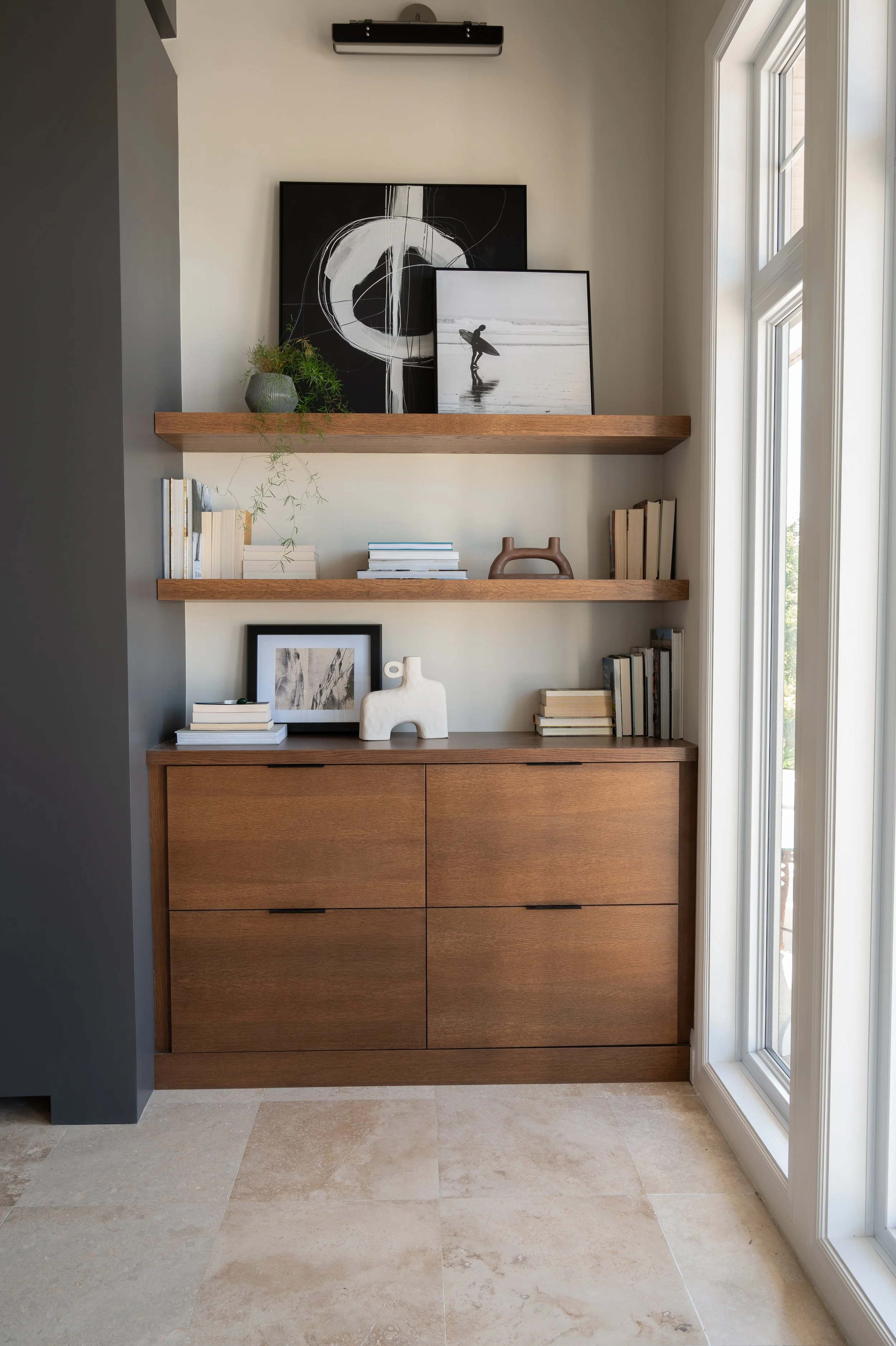 A wooden cabinet with four drawers against a wall with two floating shelves. The top shelf has two black and white framed pictures and a potted plant. The second shelf has stacked books and a decorative brown object. The cabinet has decorative object