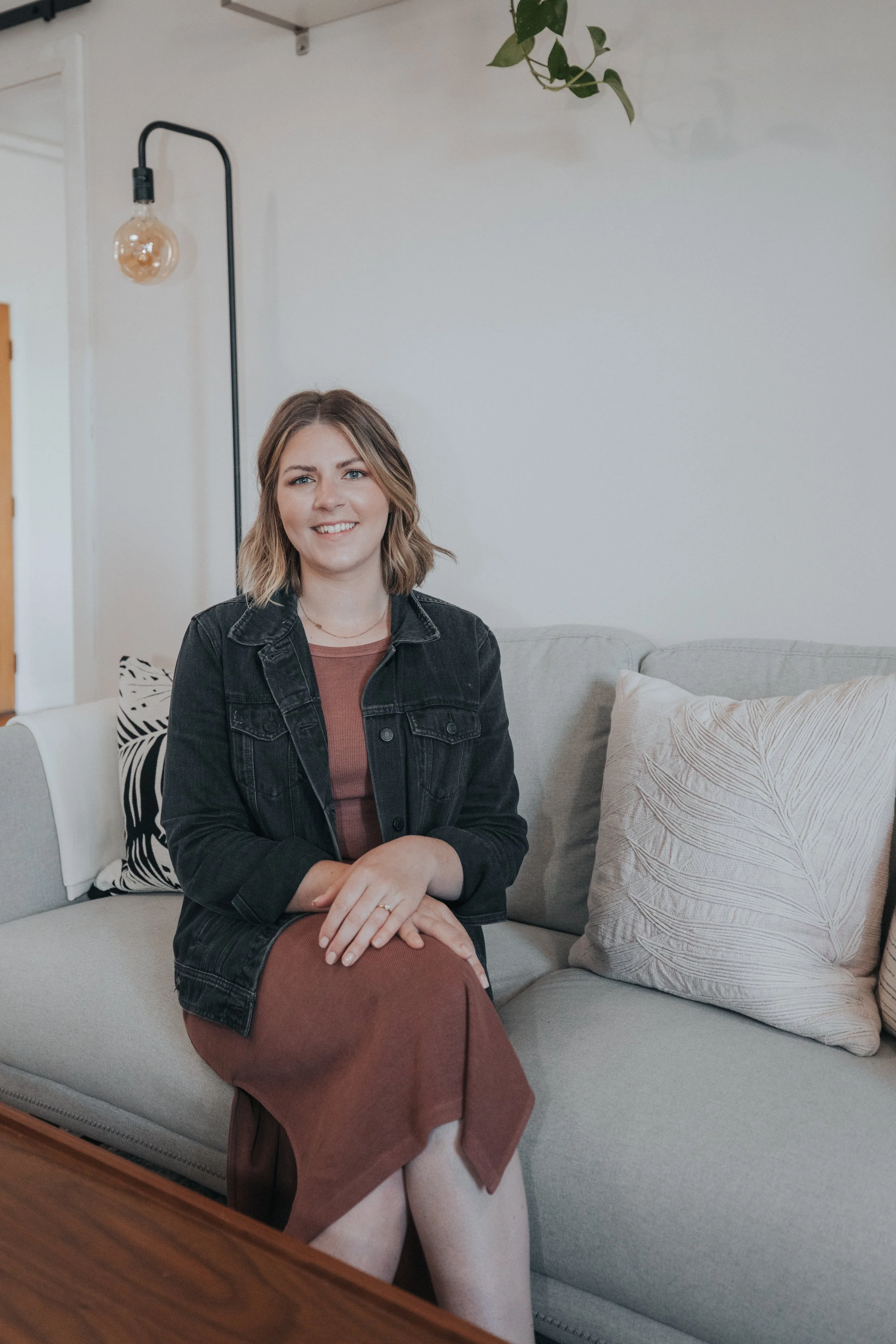 A woman sitting on a light-colored sofa in a modern, cozy living room with a black denim jacket, brown dress, and smiling at the camera.