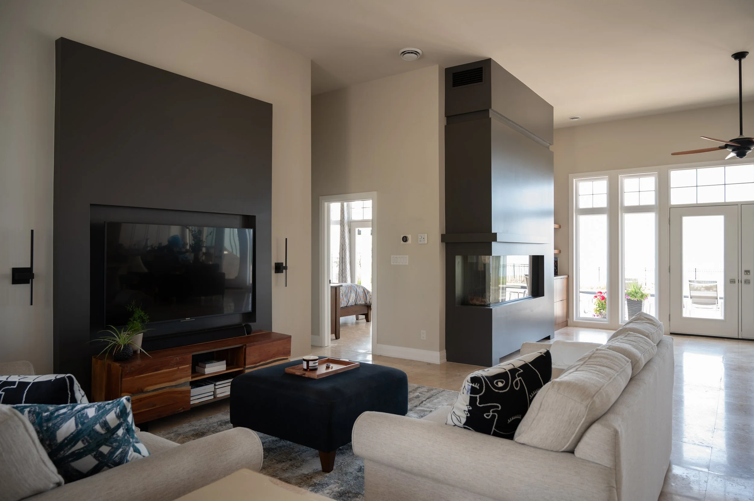 Living room with beige sofas, a black fireplace, a flat-screen TV, a wooden media console, large windows, and a view of a balcony with chairs and flowers.