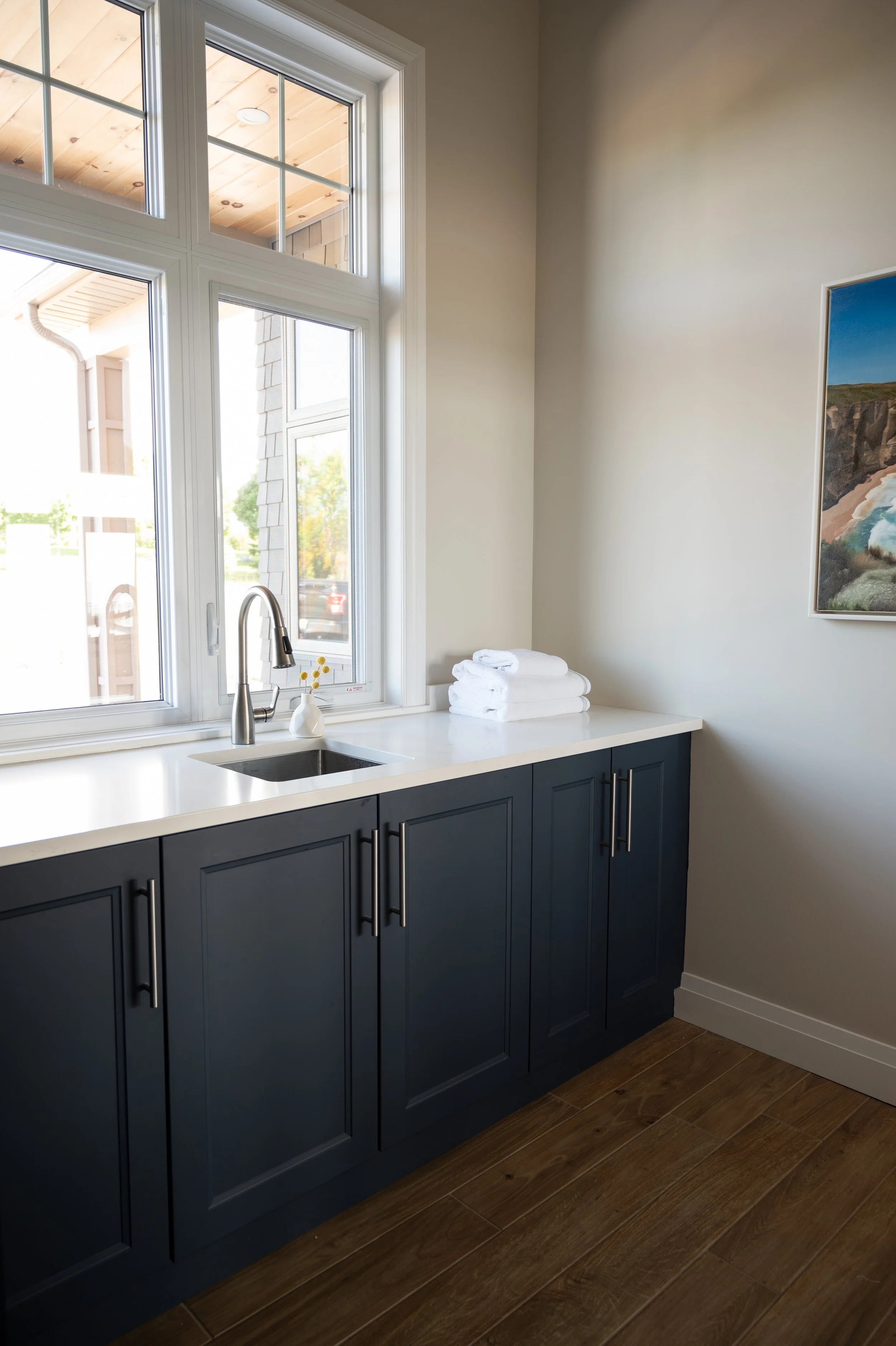 Kitchen with dark blue cabinets, white countertop, a small sink, a window above the sink, folded white towels on the counter, and a framed landscape painting on the wall.