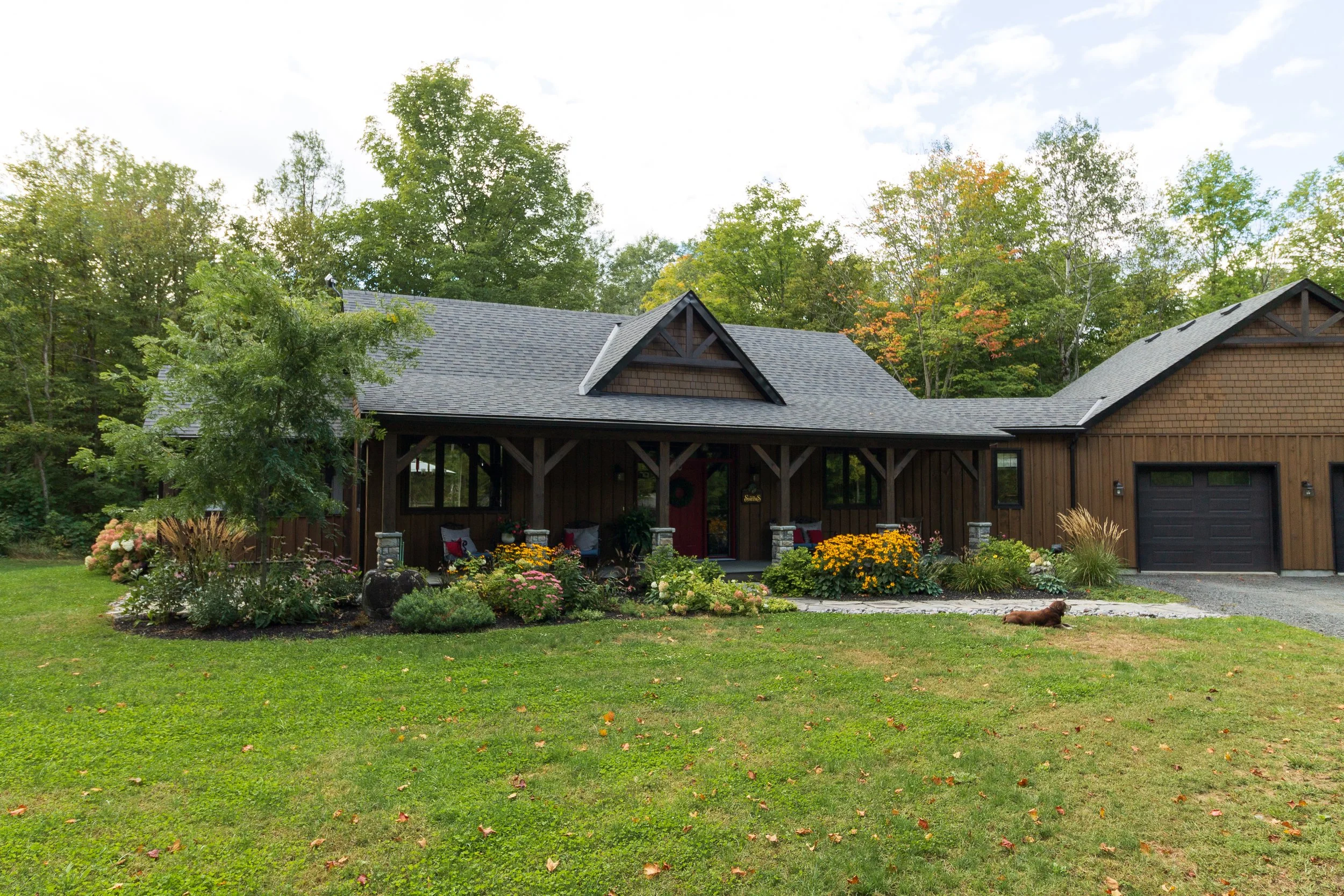 A house with a wooden exterior and gray shingle roof, surrounded by a well-maintained lawn and garden with colorful flowers, trees, and shrubbery, under a partly cloudy sky.