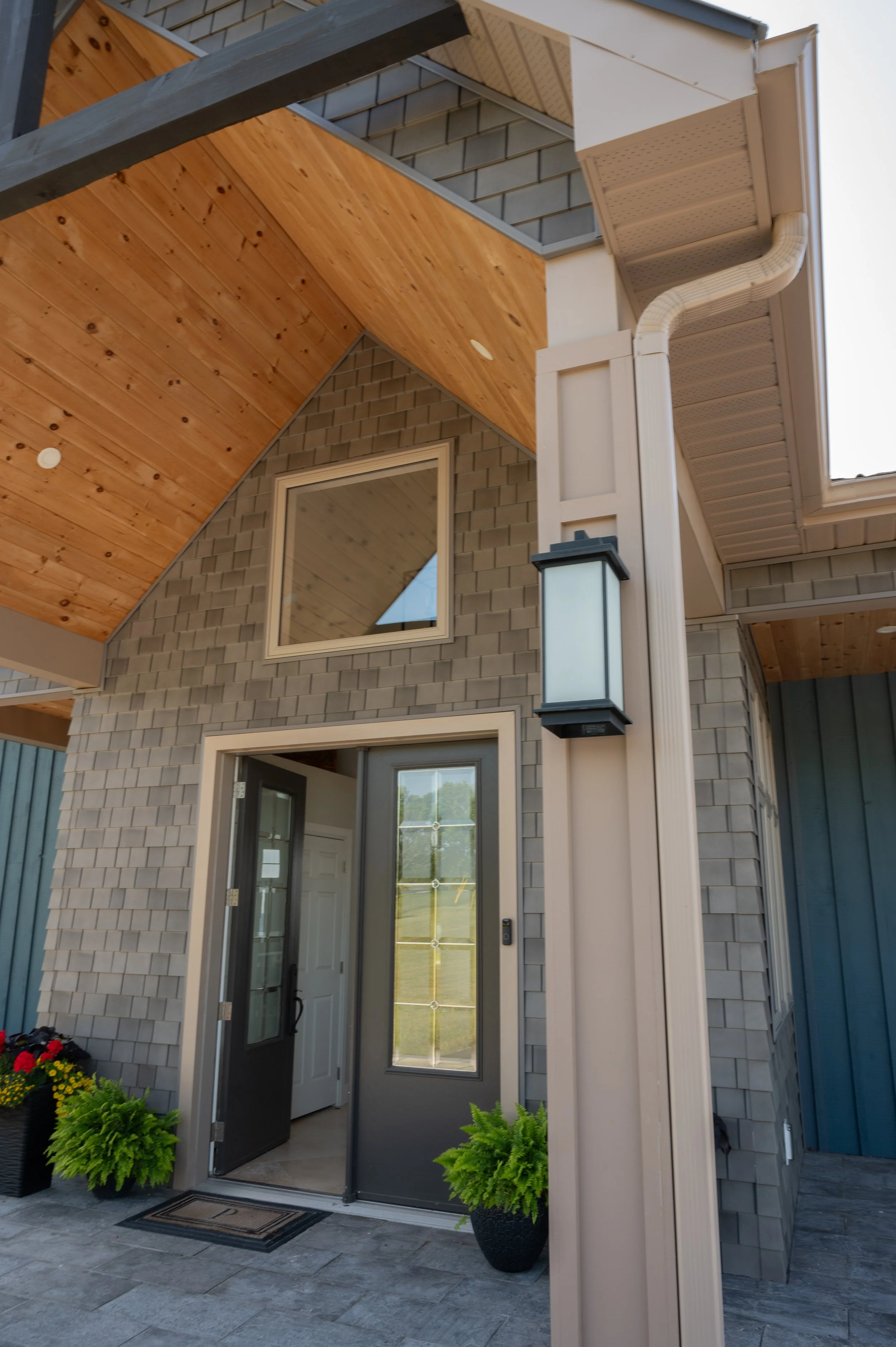 Close-up of a modern house entrance with a black door, side window, potted green plants, and exterior light fixture, with a wooden ceiling and gray brick walls.