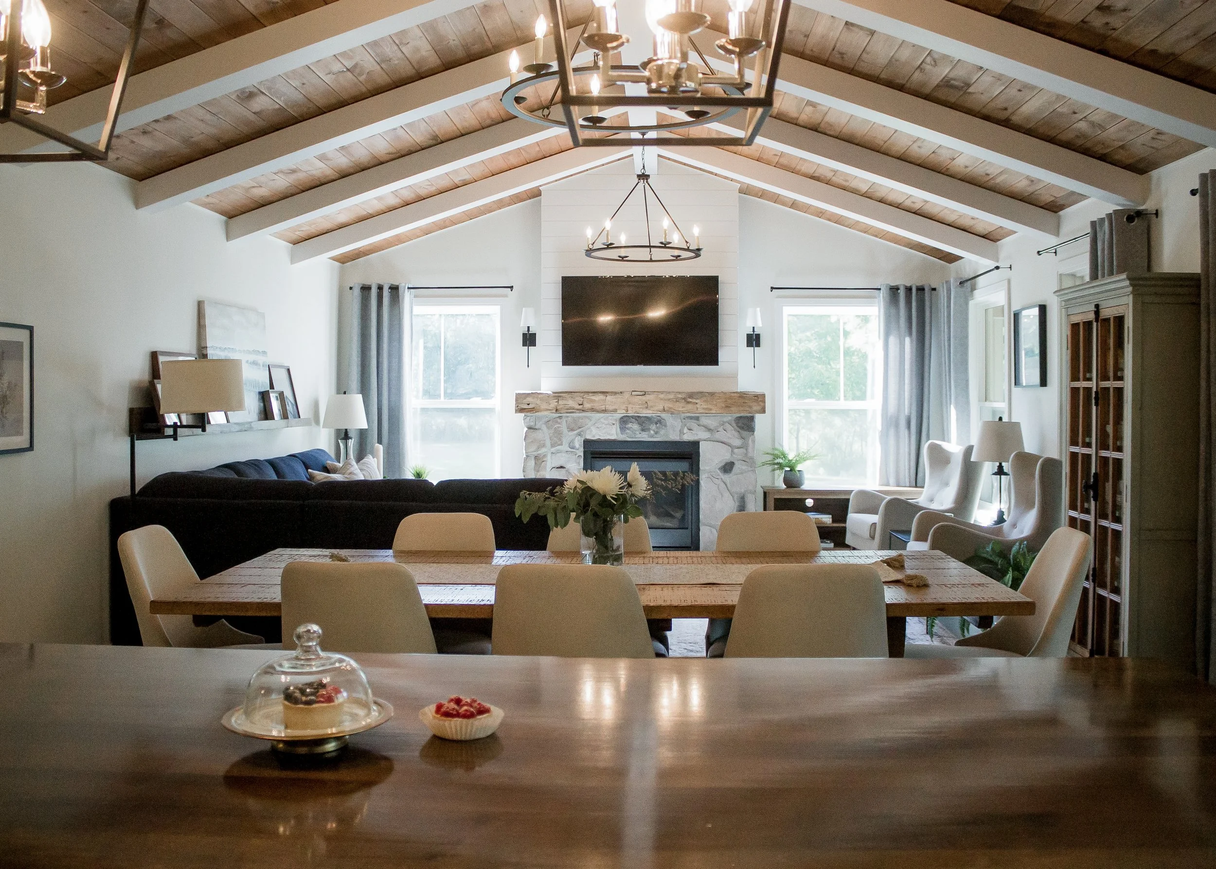 Interior of a living room with a dining table in the foreground, a stone fireplace with a mounted TV above, a black sofa, white armchairs, and large windows with gray curtains.