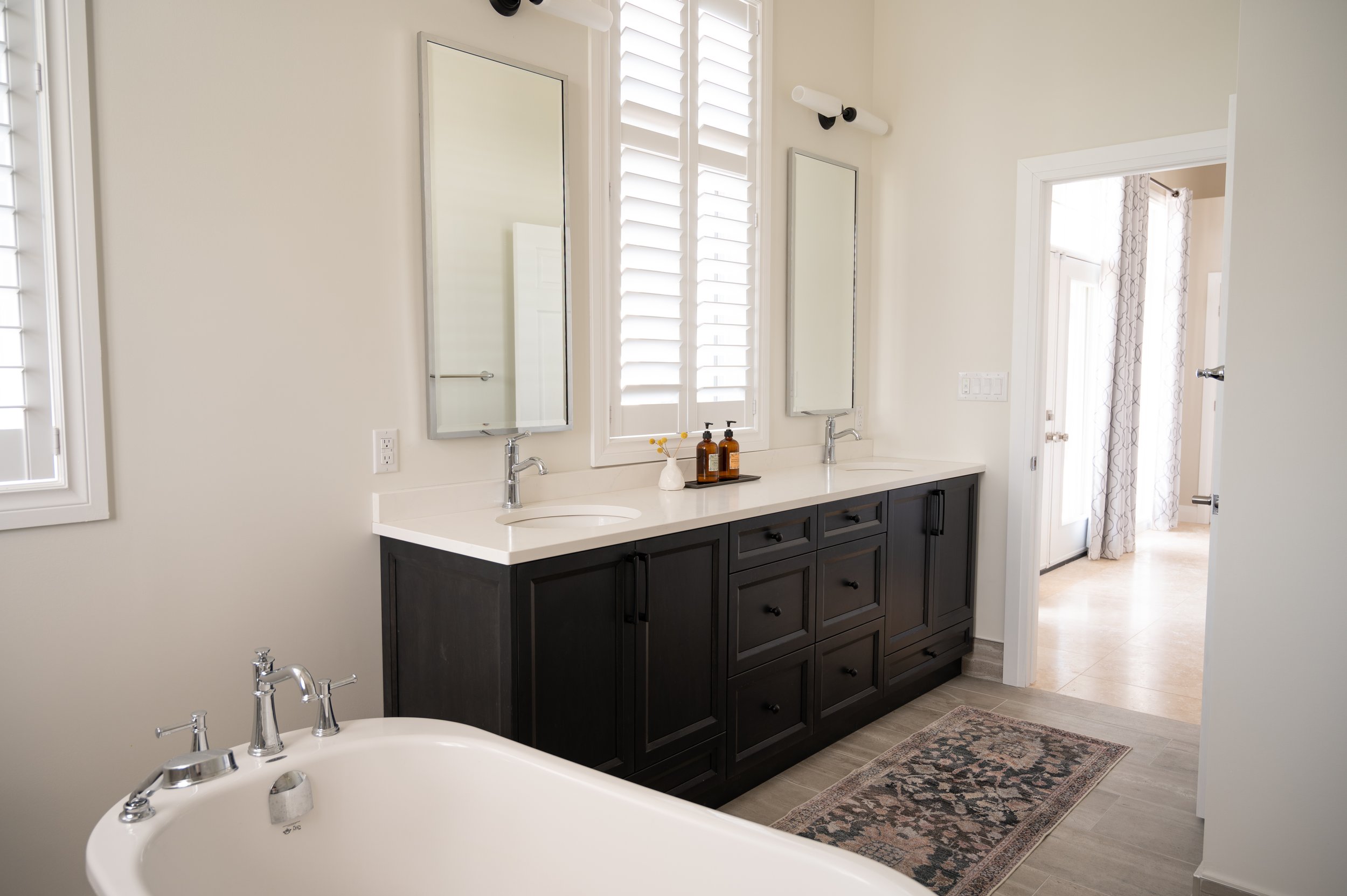 Modern bathroom with a dark wood vanity featuring two sinks,-line mirrors, and a window with white shutters. A bathtub is visible in the foreground, and a doorway leads to a bright room with patterned curtains.