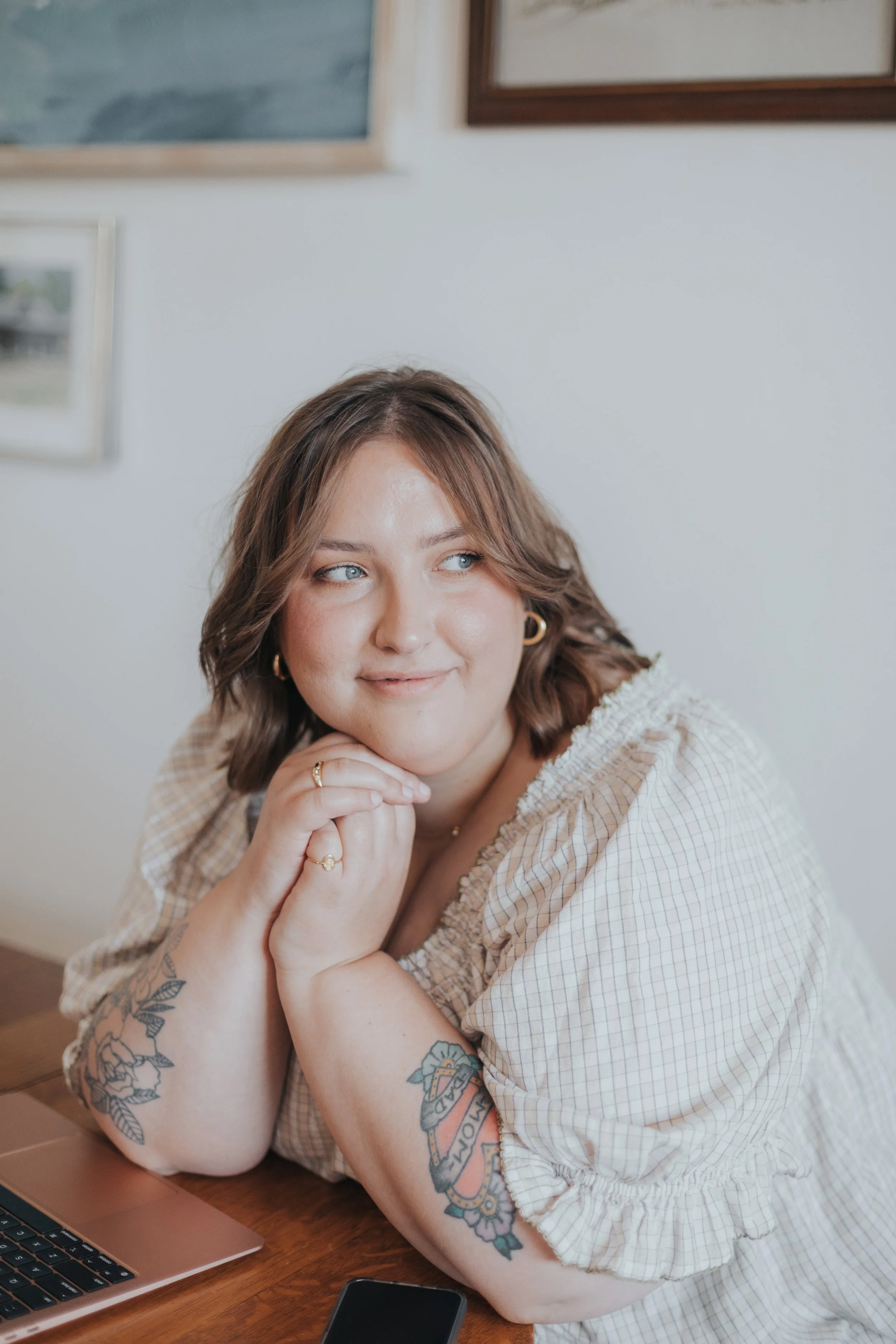 A woman with brown hair and blue eyes, wearing gold hoop earrings and tattoos on her arms, resting her chin on her hands, sitting at a wooden table with a laptop and smartphone, looking thoughtfully to the side in a well-lit room.