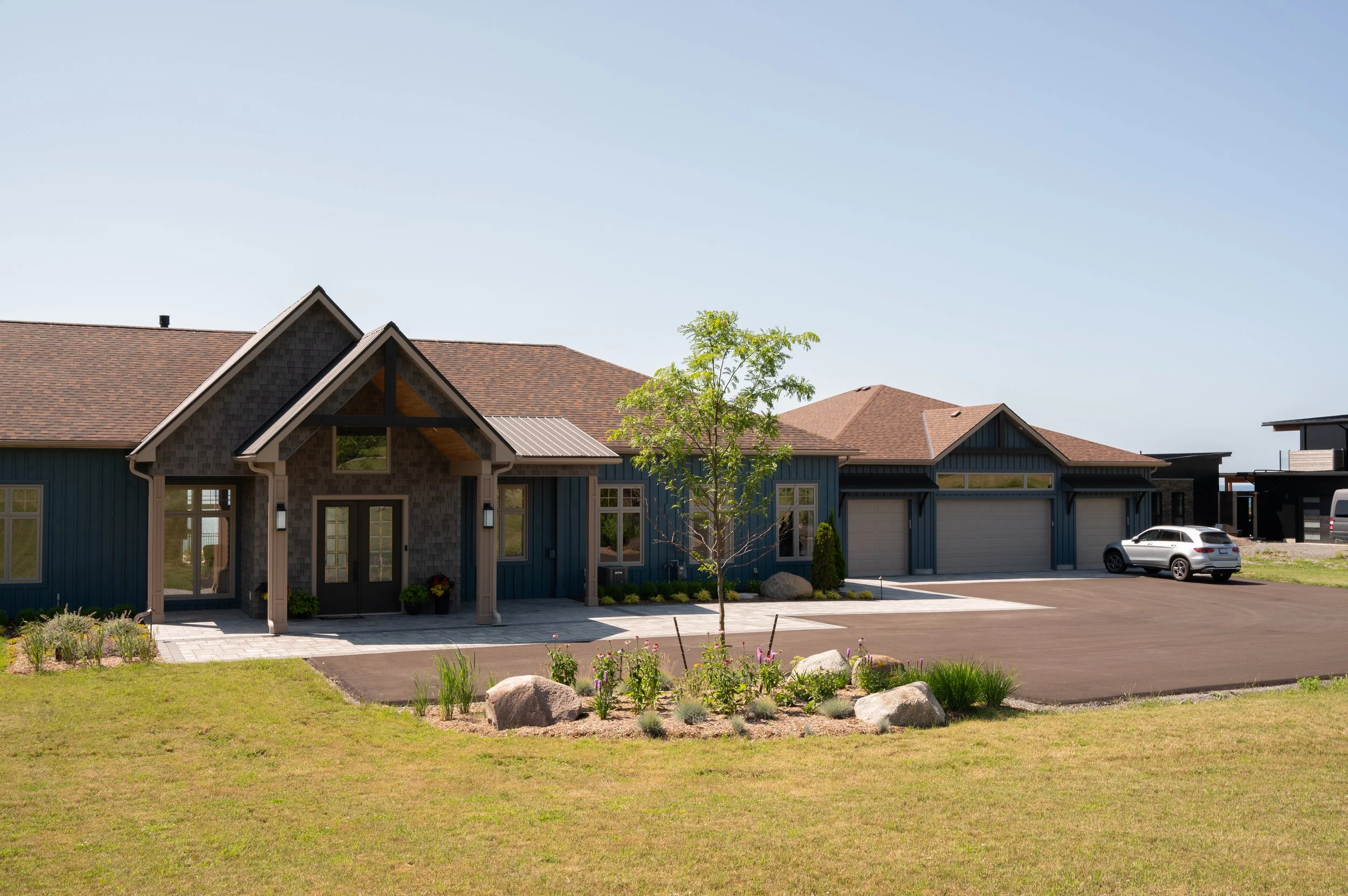 A modern suburban house with blue siding, a brown shingled roof, and a three-car garage. There is a small tree and landscaped garden with rocks and plants in the front yard. A white vehicle is parked in the driveway.