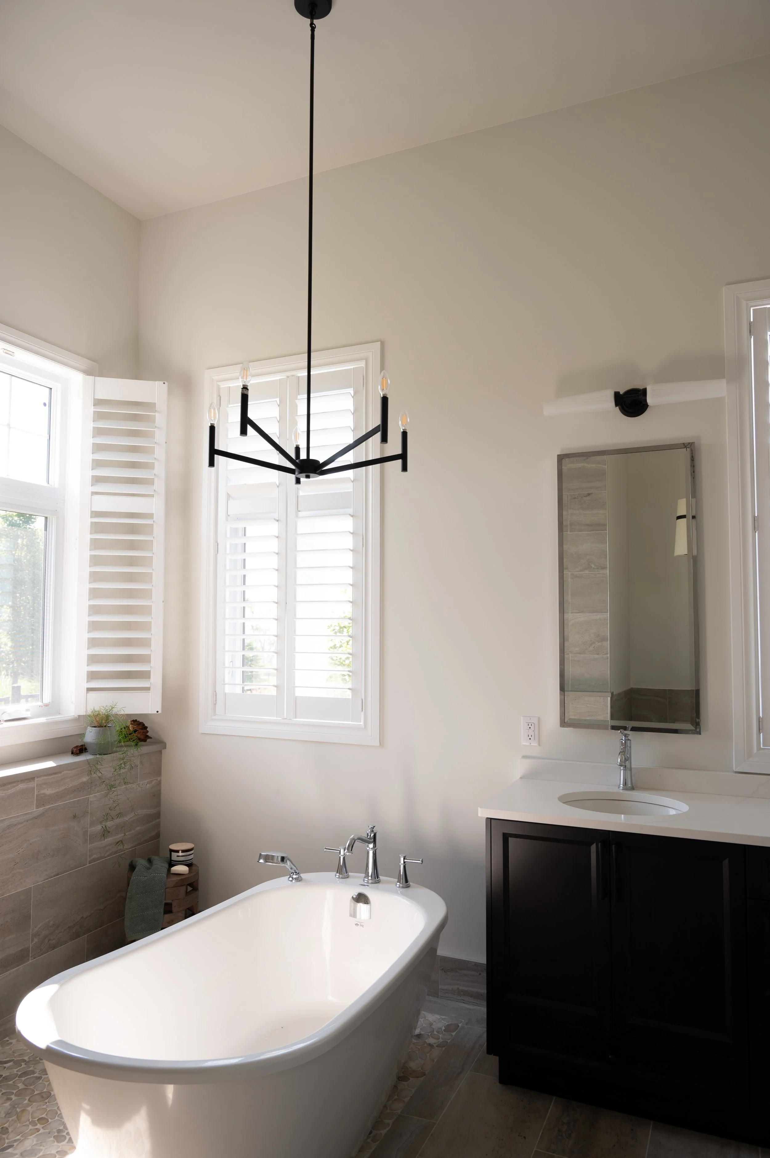 Bathroom with a white freestanding tub, black chandelier, vanity with a mirror, window with shutters, and neutral-colored tiles.