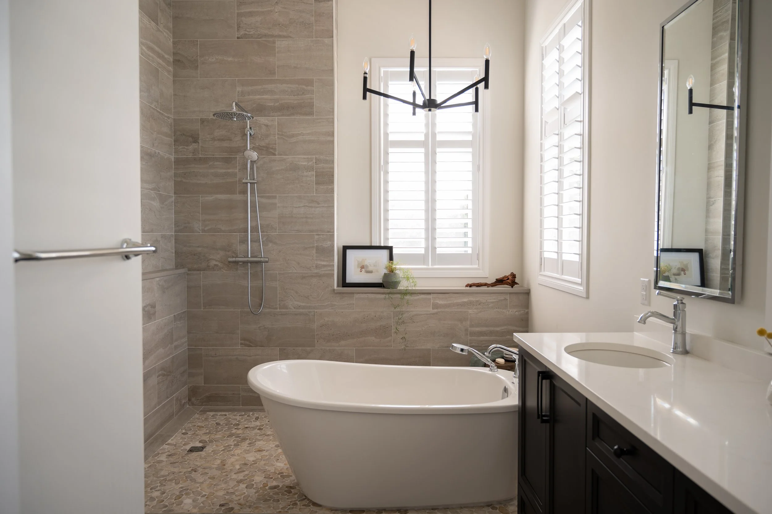Modern bathroom with bathtub, walk-in shower, vanity with mirror, window with plantation shutters, and chandelier lighting.