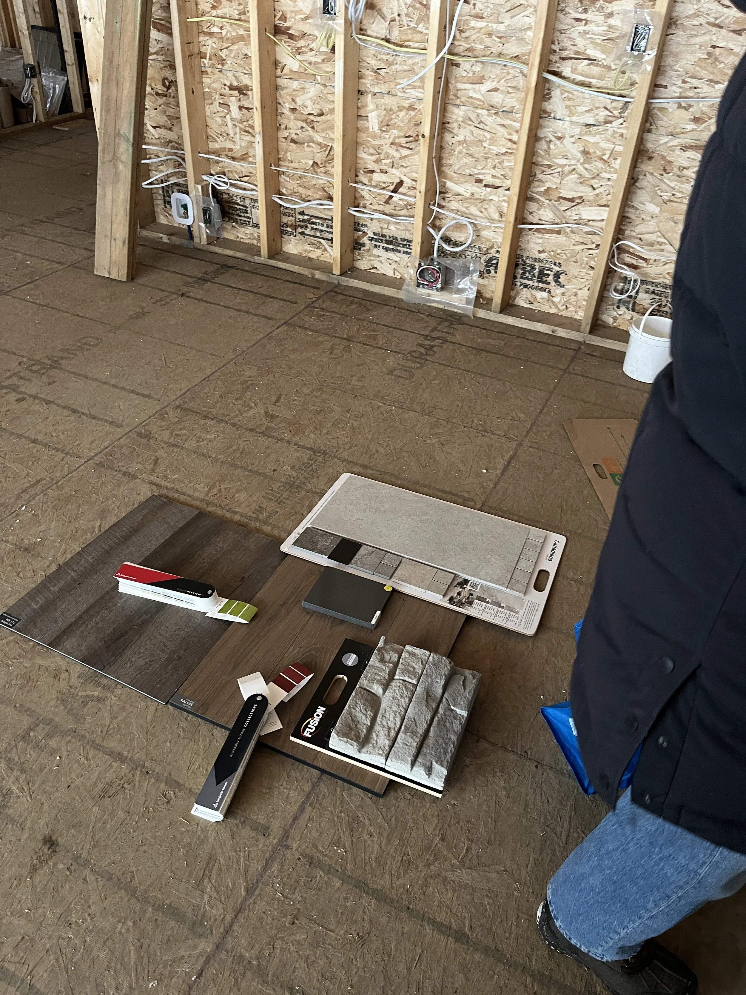 Samples of flooring and wall tiles laid out on the floor with color and material samples in an unfinished room with exposed wooden studs and electrical wiring.