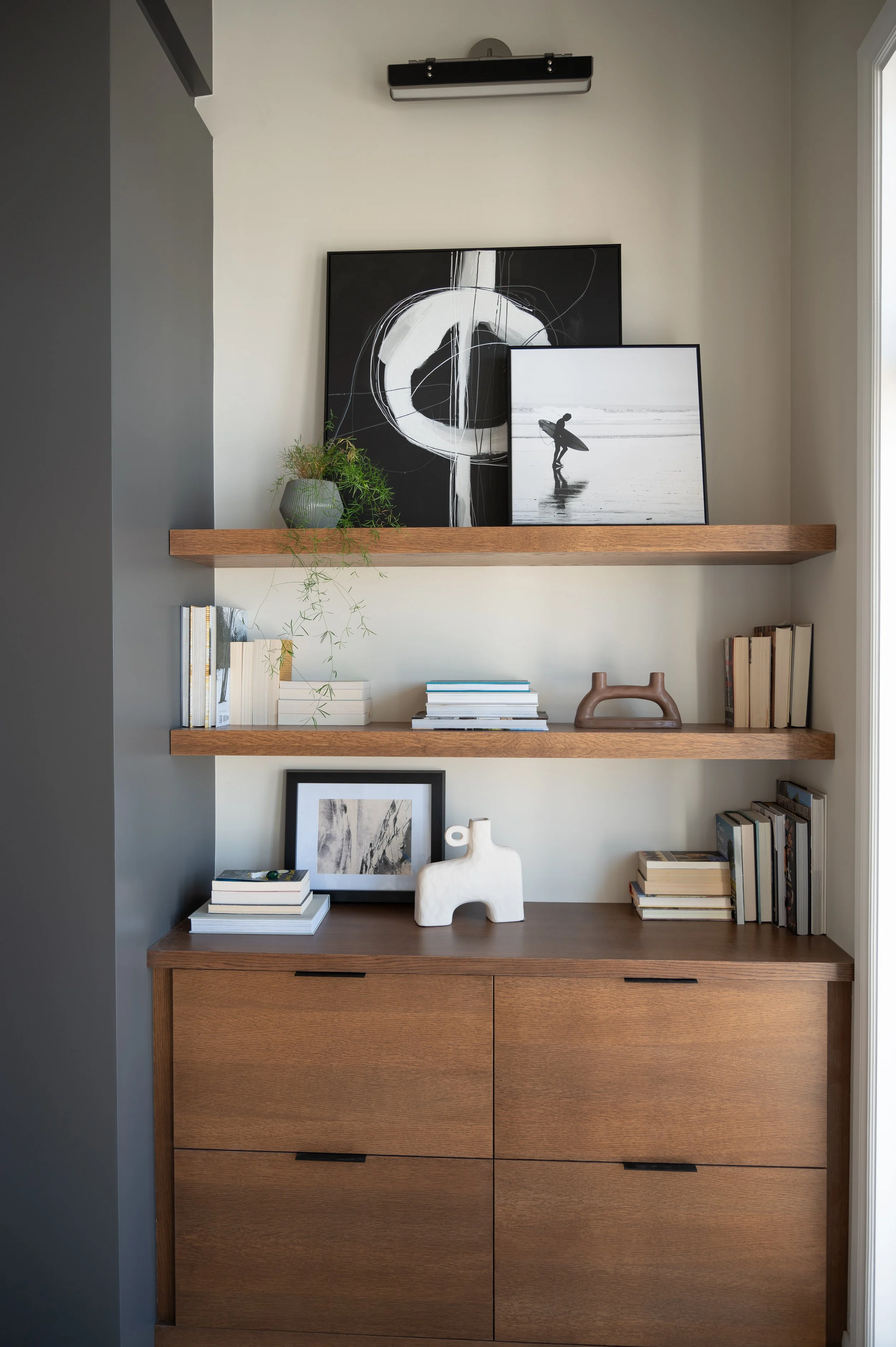A wooden sideboard with two shelves above it, decorated with books, artwork, and decorative objects, in a room with light-colored walls and a window on the right.