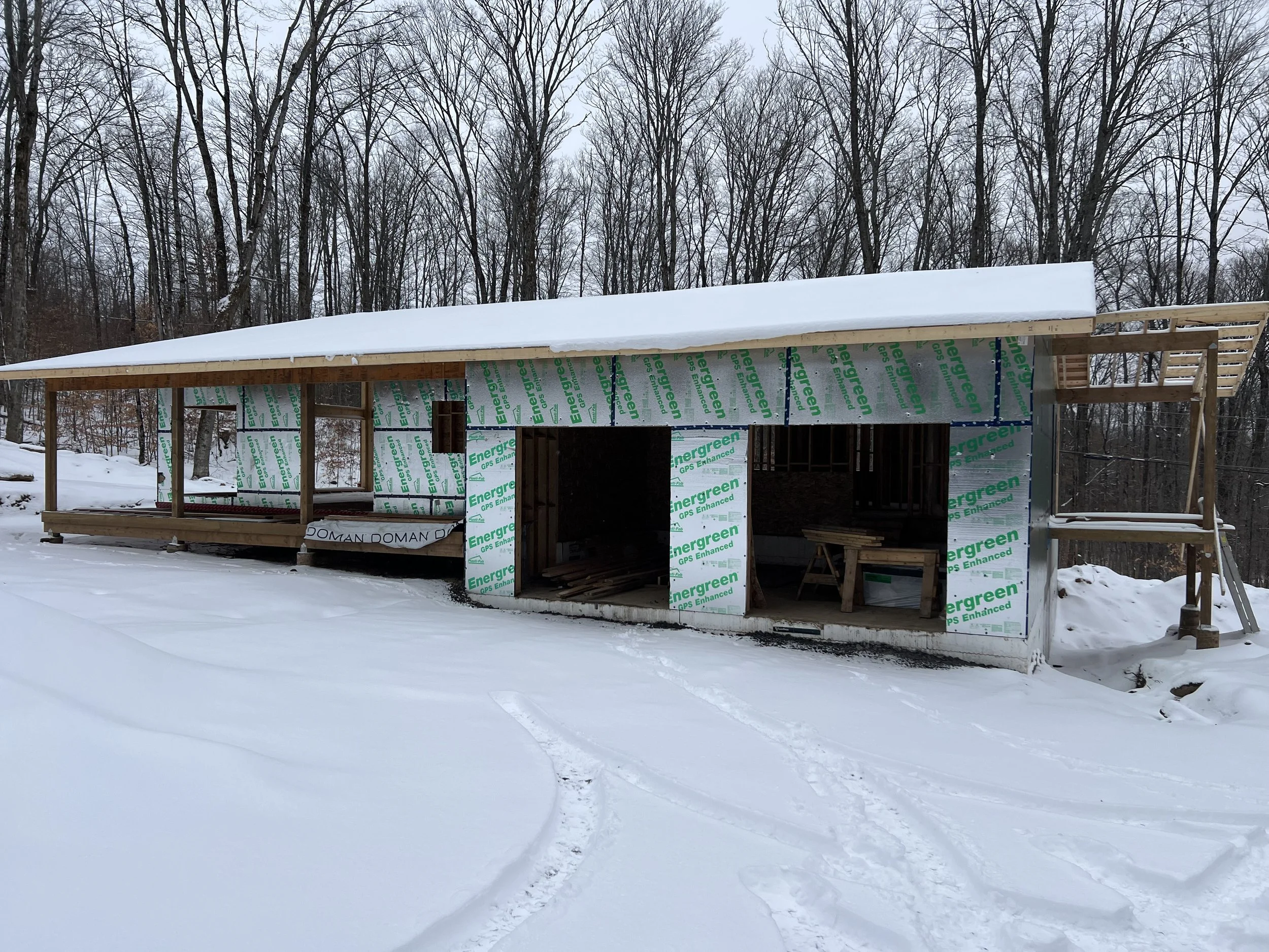 Under-construction wooden building in a snowy landscape with trees in the background.