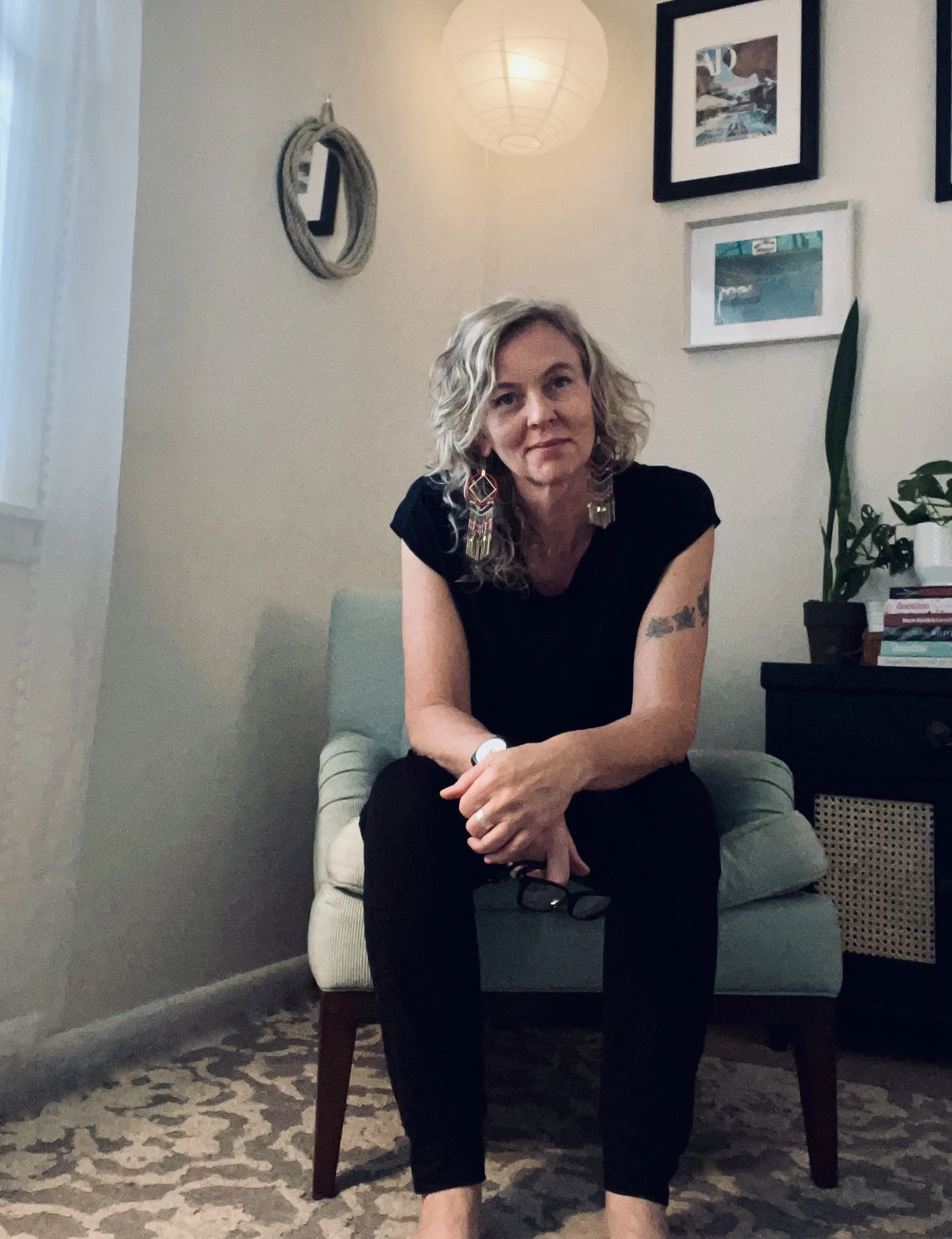 A woman with curly blonde hair sitting on a light-colored chair in a home interior, wearing a black shirt and black pants, with large colorful earrings and holding glasses. The wall behind her has framed pictures, a round decorative mirror, and a paper lantern ceiling light. A potted plant and a small stack of books are on a black side table.