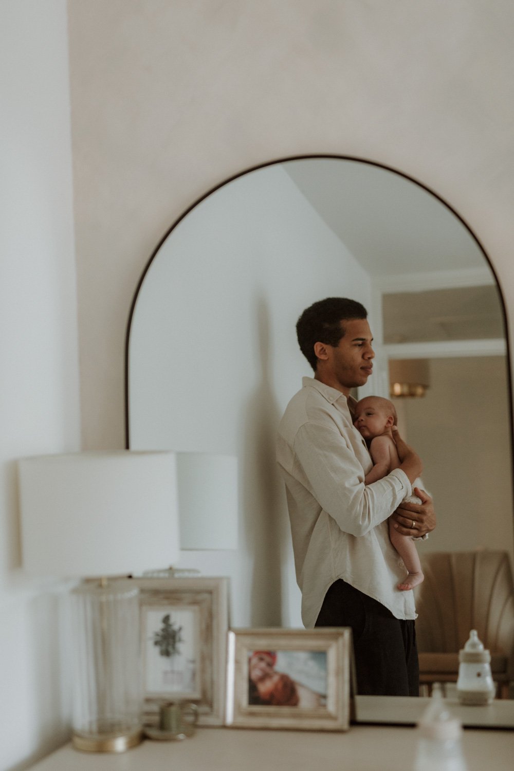 Father standing by the bedroom wall holding his newborn on his chest, a quiet, reflective moment in a natural at-home session