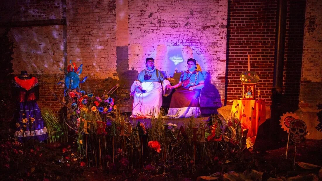 Mexican Day of the Dead altar with two mannequins dressed as traditional figures, surrounded by colorful flowers, skeleton decorations, and festive objects, set against a brick wall.
