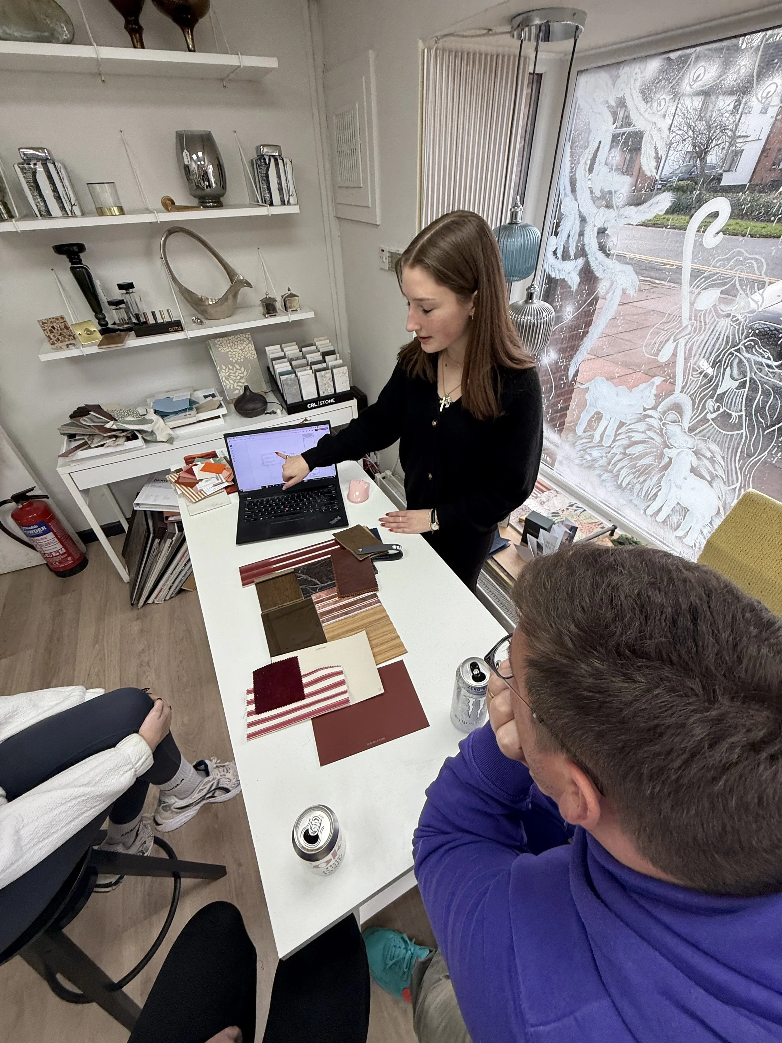 A woman in black top is pointing at her laptop screen on a white desk surrounded by sample fabric swatches. She is in a room with shelves holding various decorative items, and a large window with frosted designs and a street view outside. Two people are seated at the table, one woman in a white jacket and another man in a blue hoodie.
