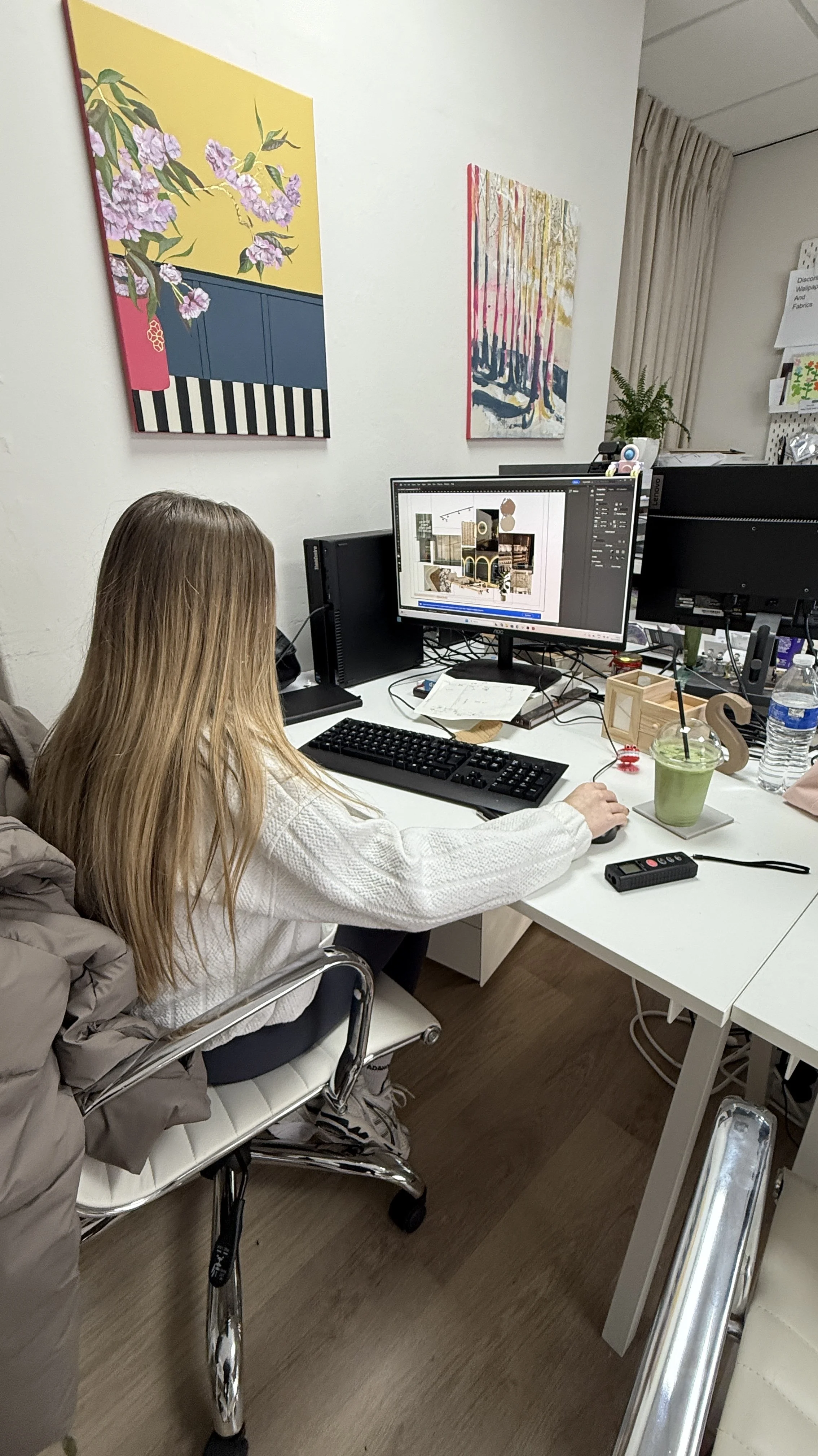 A woman with long light brown hair sitting at a white desk, working on a computer with a design software open. The desk has drinks, a remote control, and papers. Behind her are two colorful abstract paintings on the wall, and a potted plant is nearby.