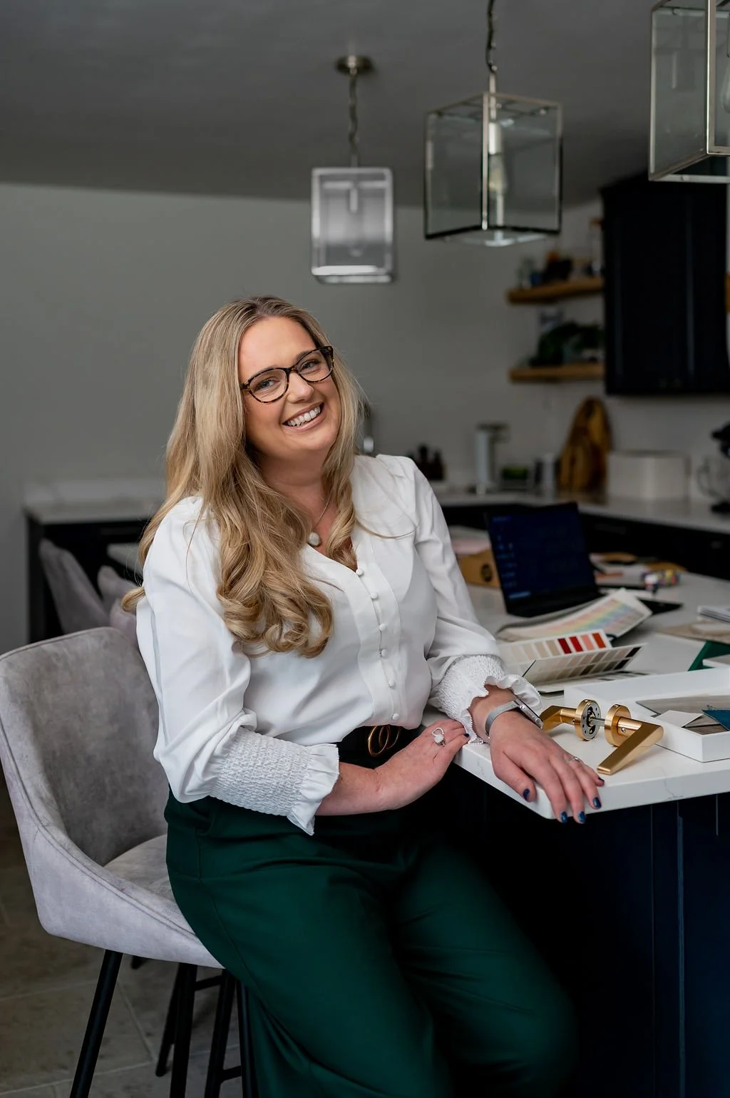 A woman with glasses and long blonde hair smiling while sitting at a kitchen counter with various decor and work items, including a laptop and color swatches.