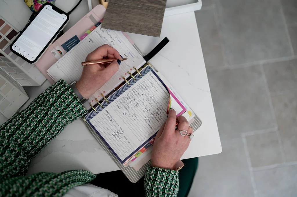 A person writing in a planner on a desk surrounded by stationery, a smartphone, and some papers.