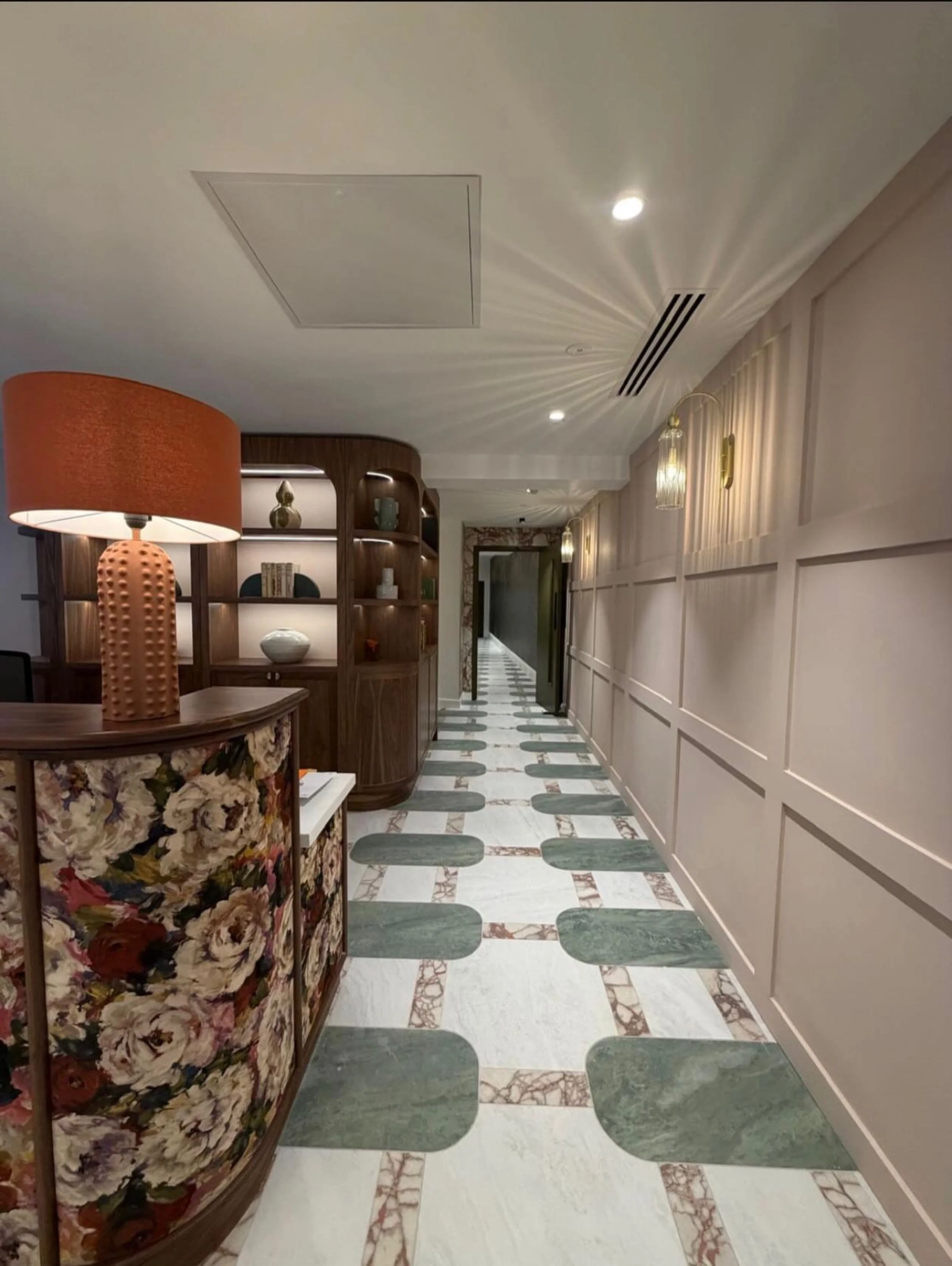 Interior hallway with a marble tiled floor, wooden shelving on the left with decorative objects, and beige paneled walls with hanging light fixtures on the right side.