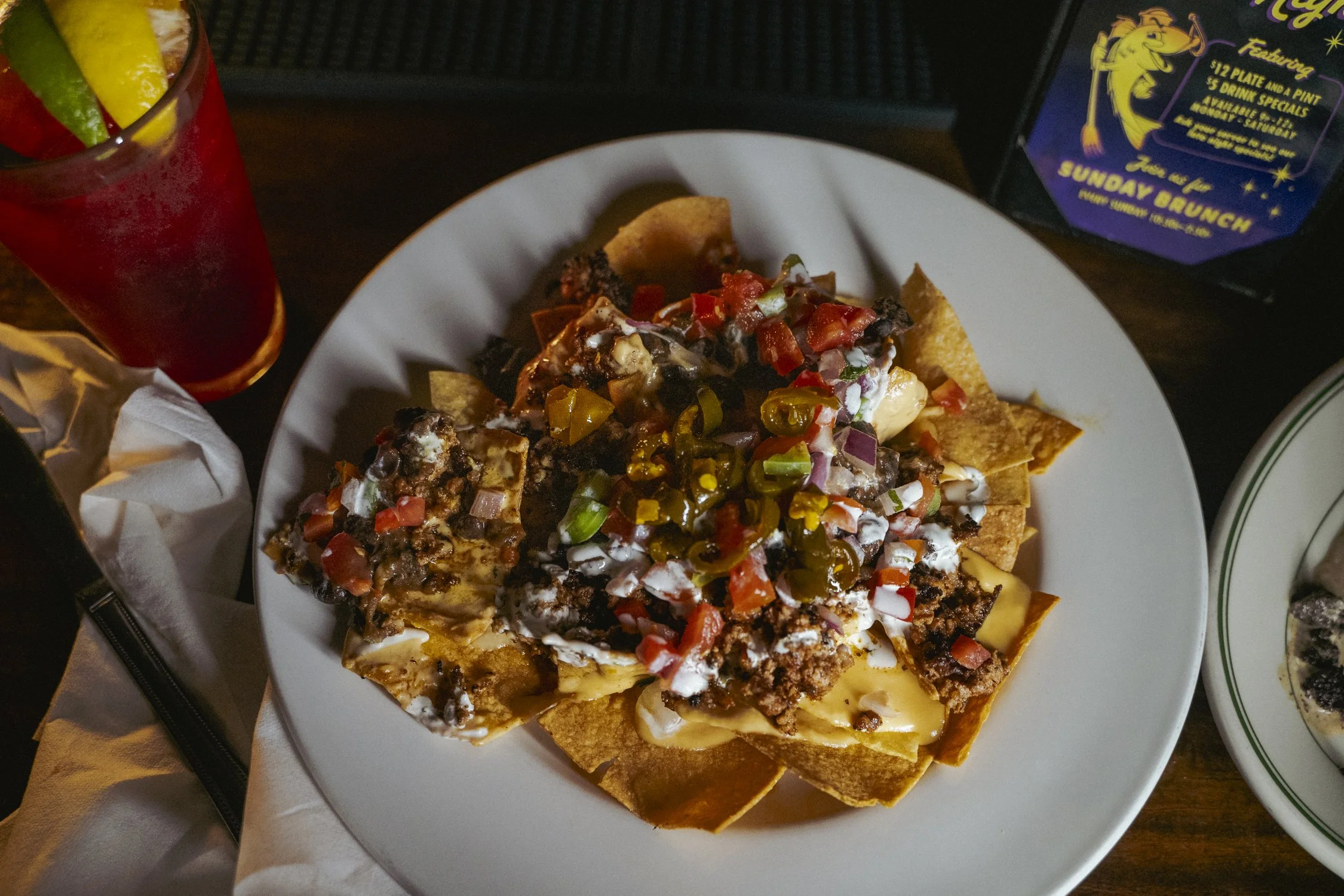Close-up of a hand placing a tortilla chip with melted cheese, on a plate of nachos topped with ground beef, diced tomatoes, chopped onions, sliced jalapeños, black beans, and sour cream.