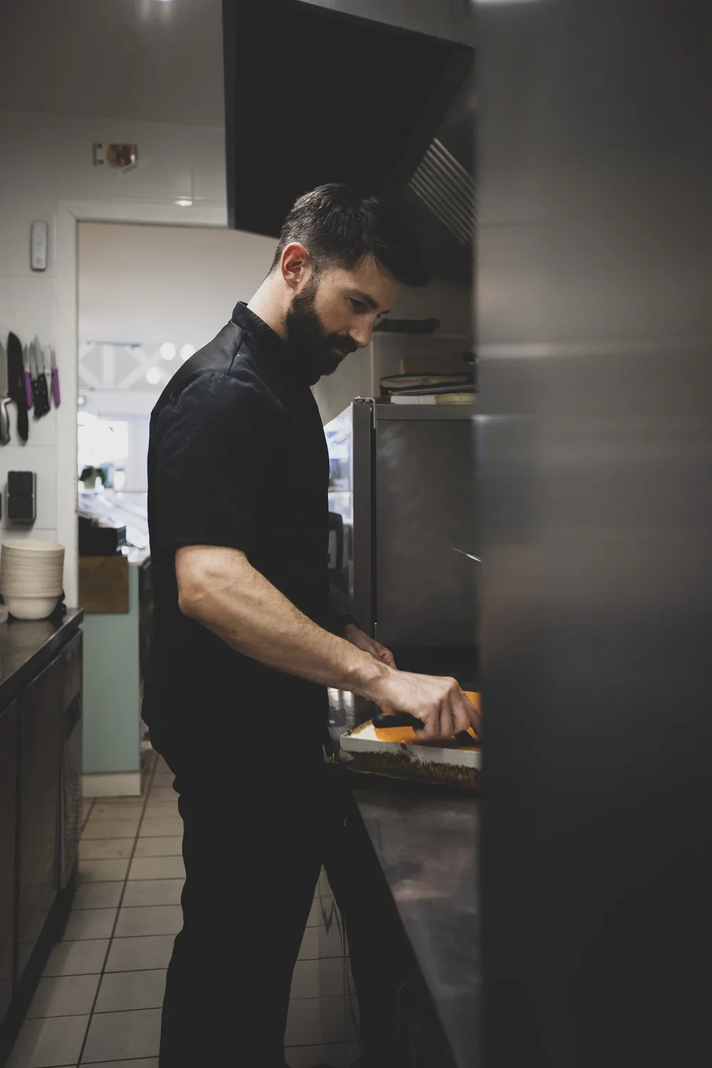 Un homme avec une barbe, portant une chemise noire, coupe quelque chose dans une cuisine professionnelle.