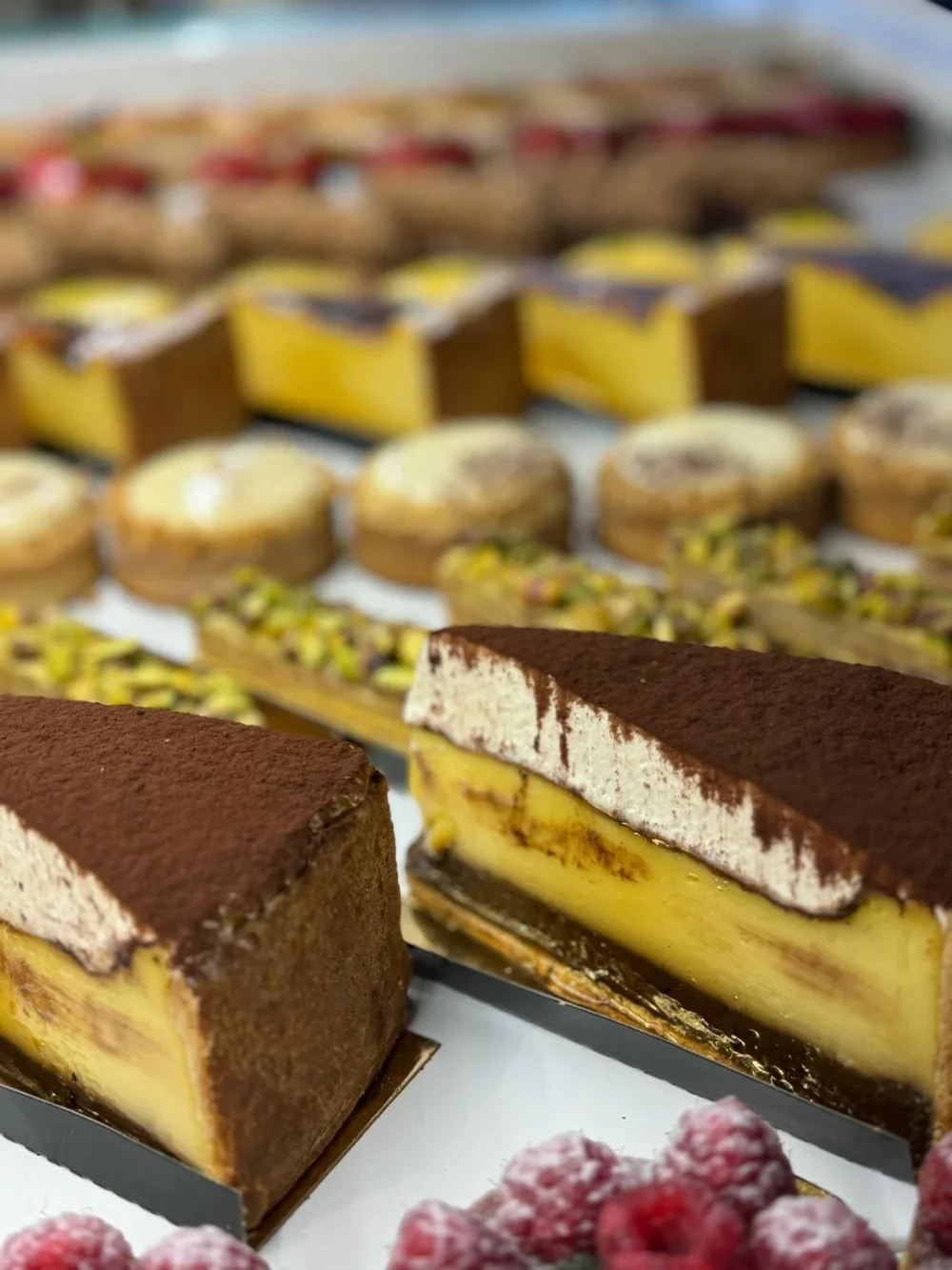 Gros plan d'un gâteau au tiramisu avec une couche de cacao en poudre et une couche de mascarpone, entouré d'autres desserts comme des tartes, des cheesecakes et des éclairs, en vitrine de pâtisserie.