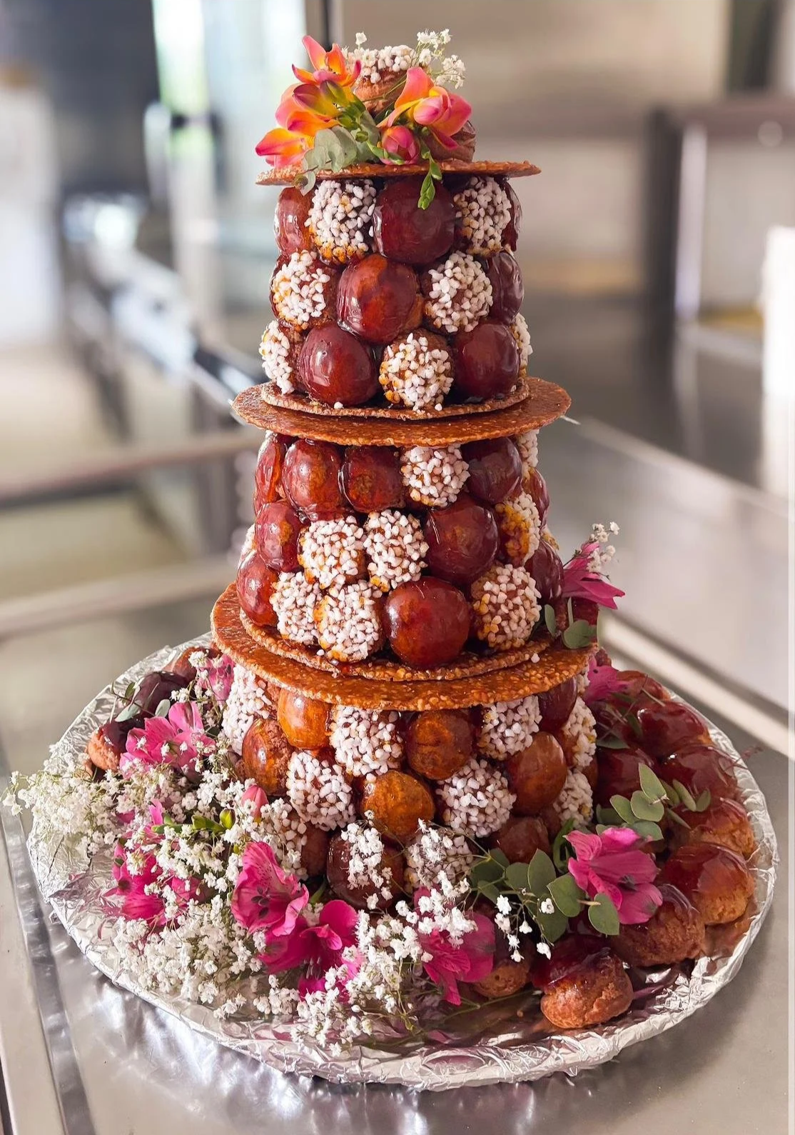 Un gâteau en forme de tour, composé de choux confectionnés avec des chapeaux en caramel et décoré de fleurs colorées, de sucre pailleté blanc, sur une plaque en aluminium avec des fleurs roses et blanches autour.