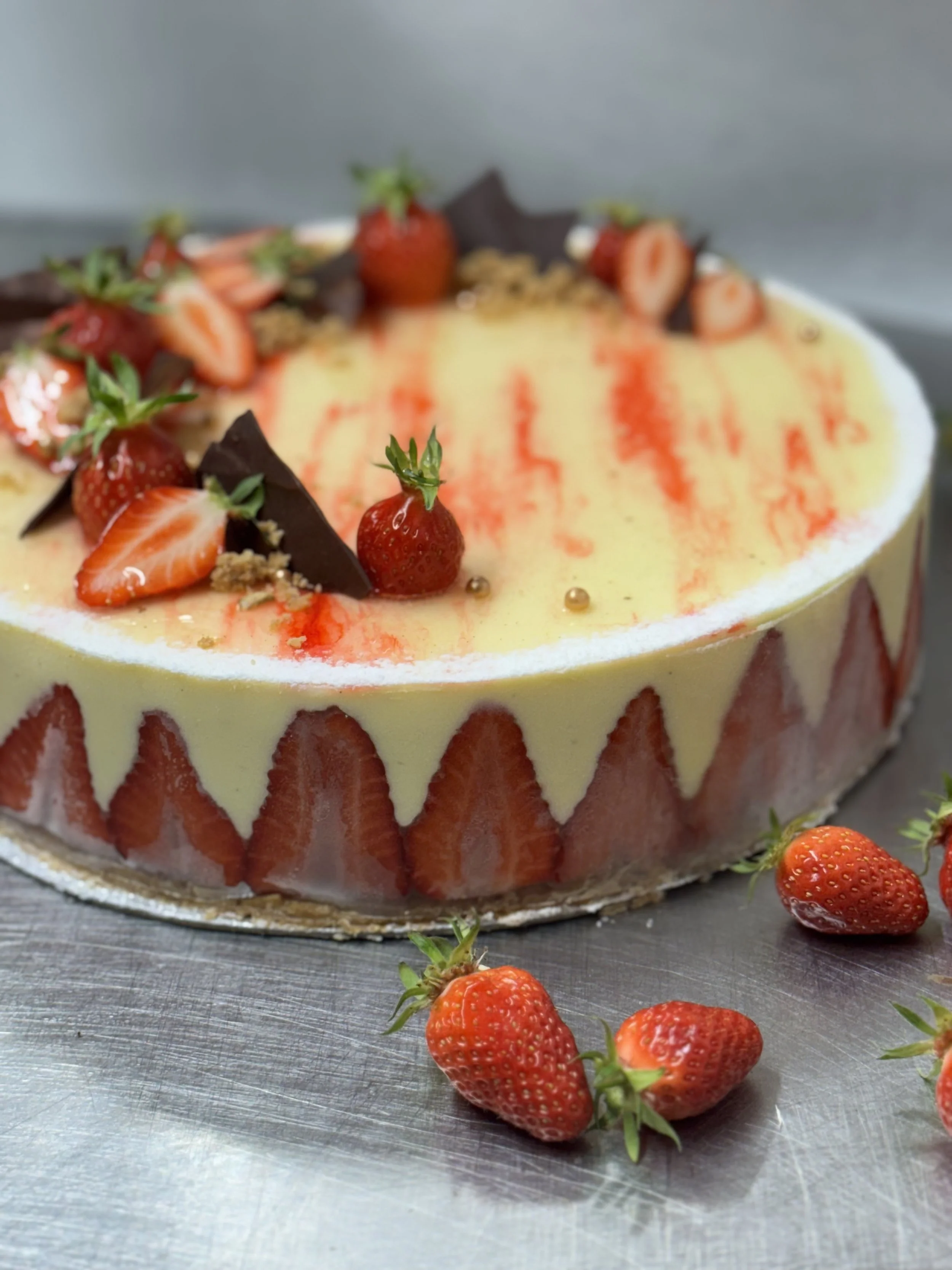 Gâteau aux fraises décoré avec des morceaux de fraises, des feuilles vertes et des morceaux de chocolat, placé sur une surface en métal.