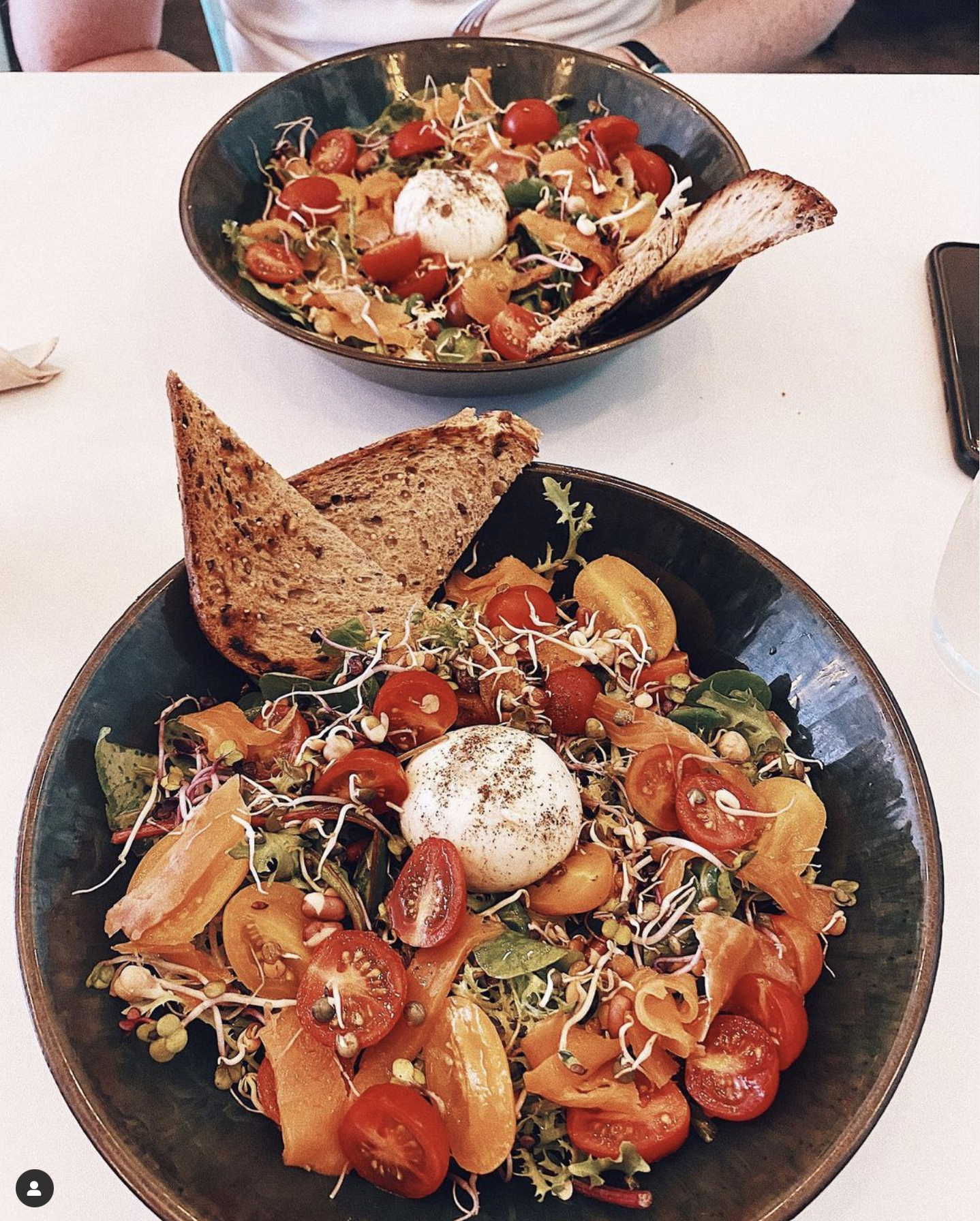 Salades avec tomates, œuf, germes, pain grillé, servies dans des bols noirs sur une table blanche.