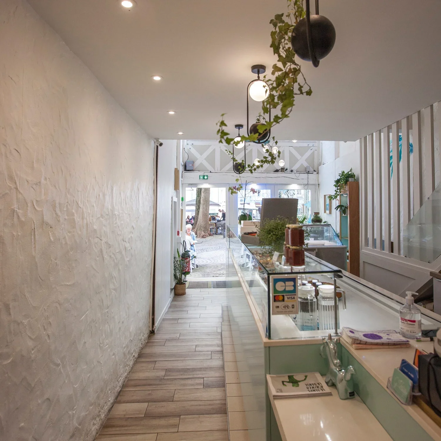 Intérieur d'un café ou une boutique avec comptoir en verre, plantes en décoration, porte d'entrée donnant à l'extérieur, et table avec clients dehors.