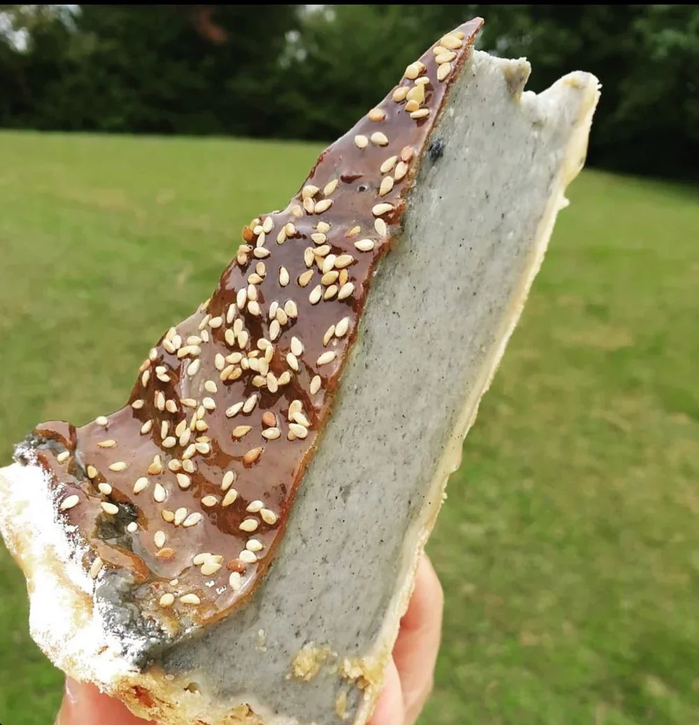 Gâteau en forme de tranche de tôle rouillée, recouvert de glaçage au chocolat et saupoudré de graines de sésame, tenu en main. Fond vert extérieur.