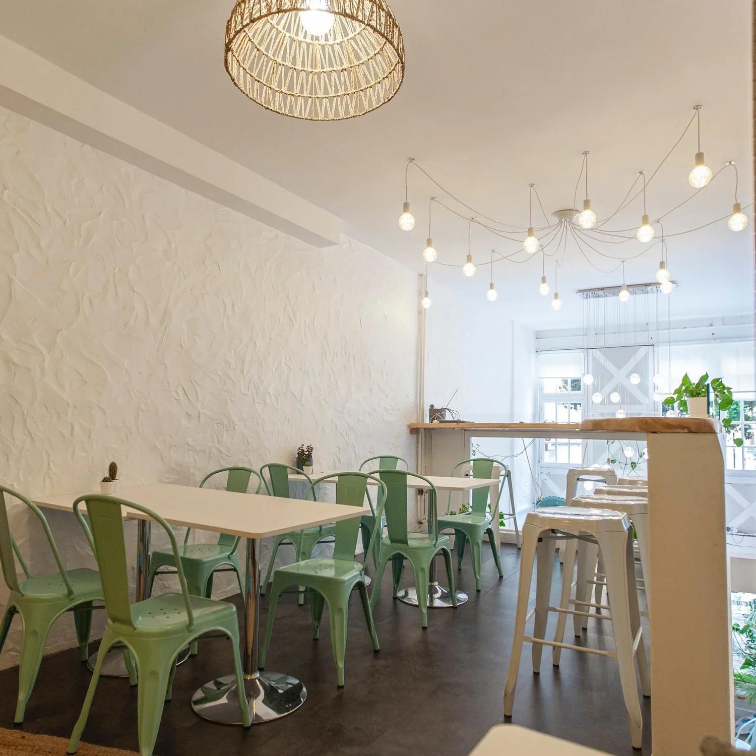 Intérieur d'un café lumineux avec des tables vertes, des chaises en métal vert et des tabourets blancs, une grande lampe suspendue en rotin, et des ampoules décoratives suspendues au plafond, avec des plantes en arrière-plan.