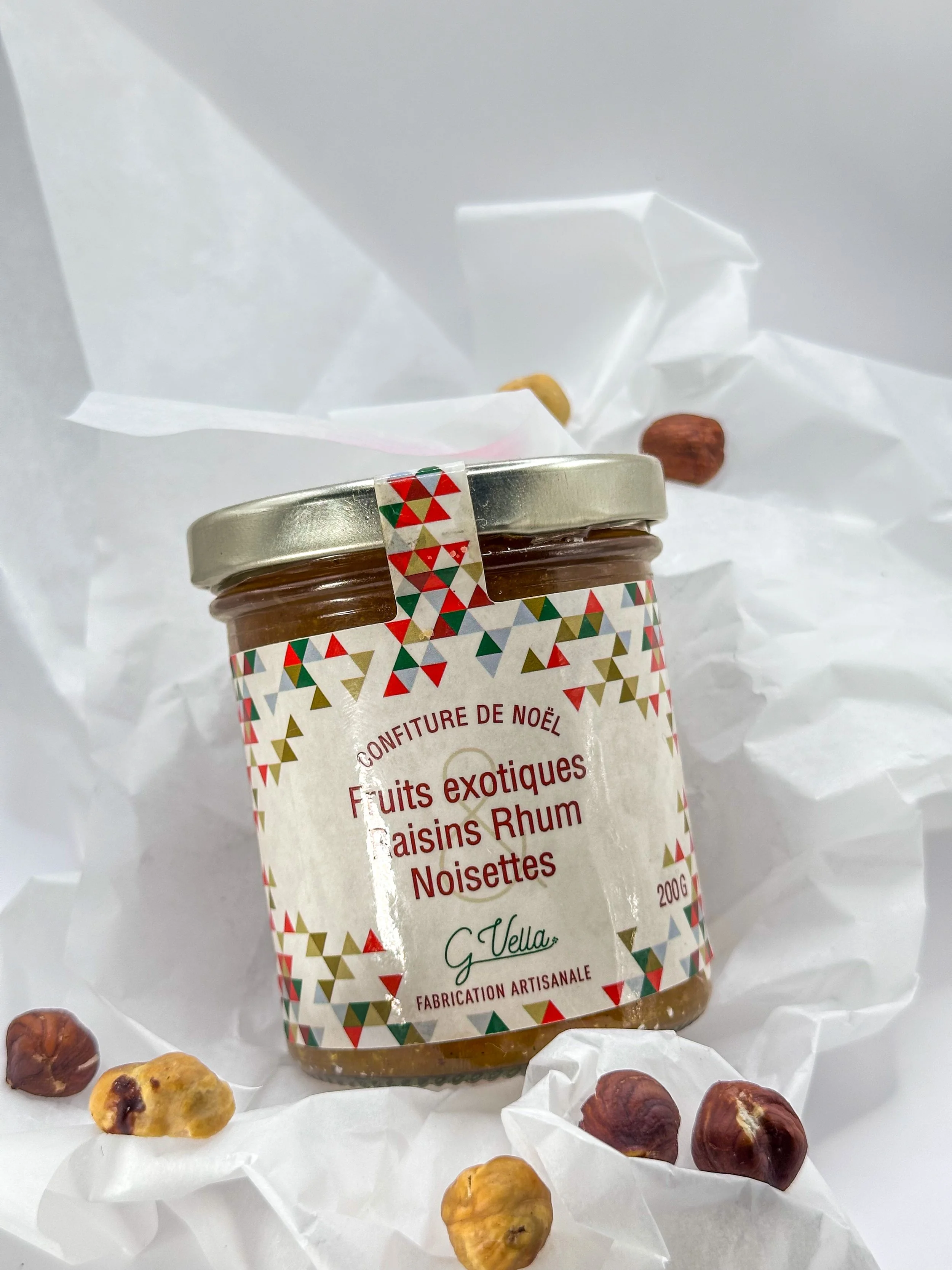 Un pot de confiture aux fruits exotiques, raisins, rhum et noisettes, entouré de noisettes et raisins secs sur du papier blanc.