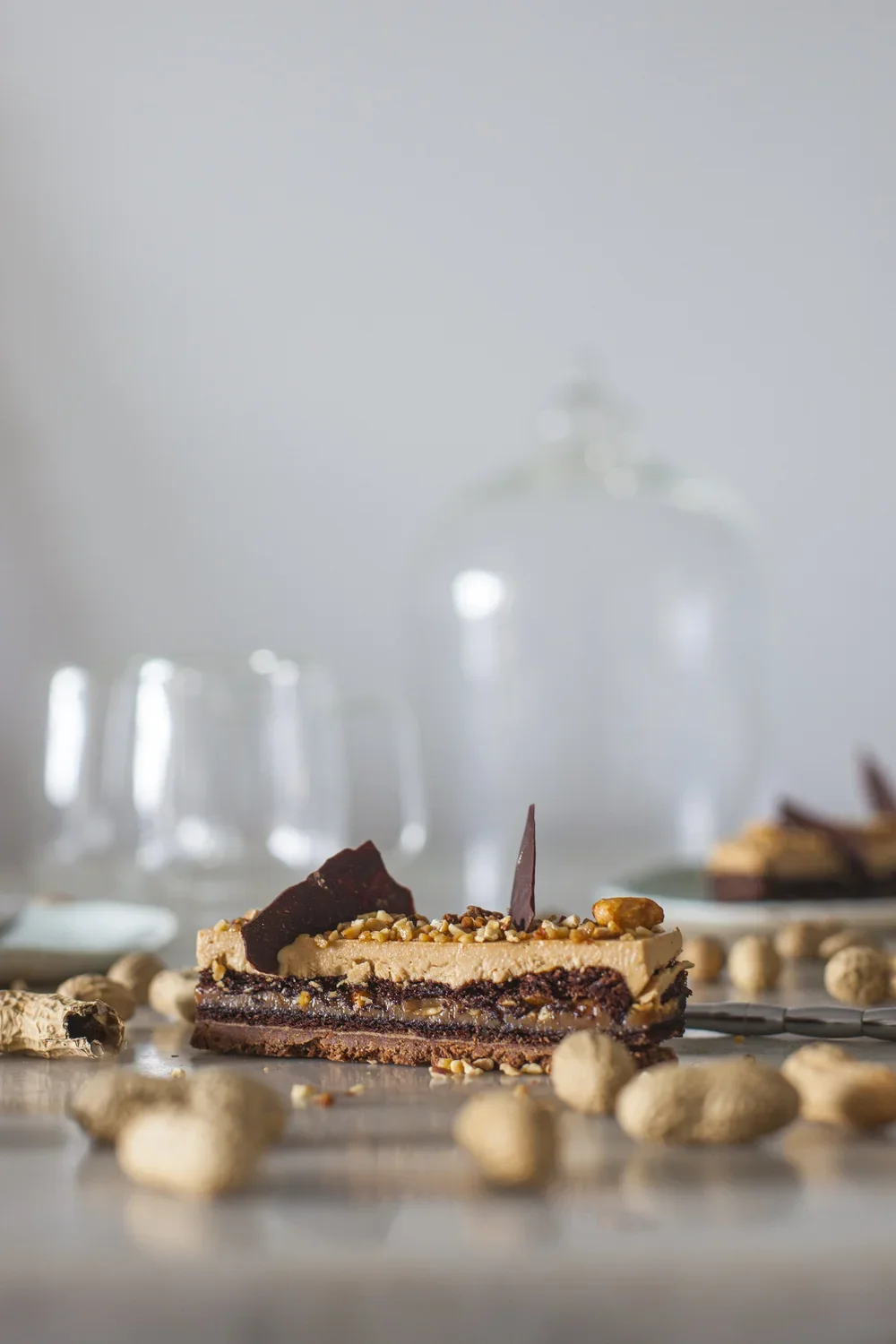 Une part de gâteau au chocolat et café, décorée de noix et de chocolat, avec des noix entières éparpillées devant, sur une surface en bois, avec des verres à vin flous en arrière-plan.