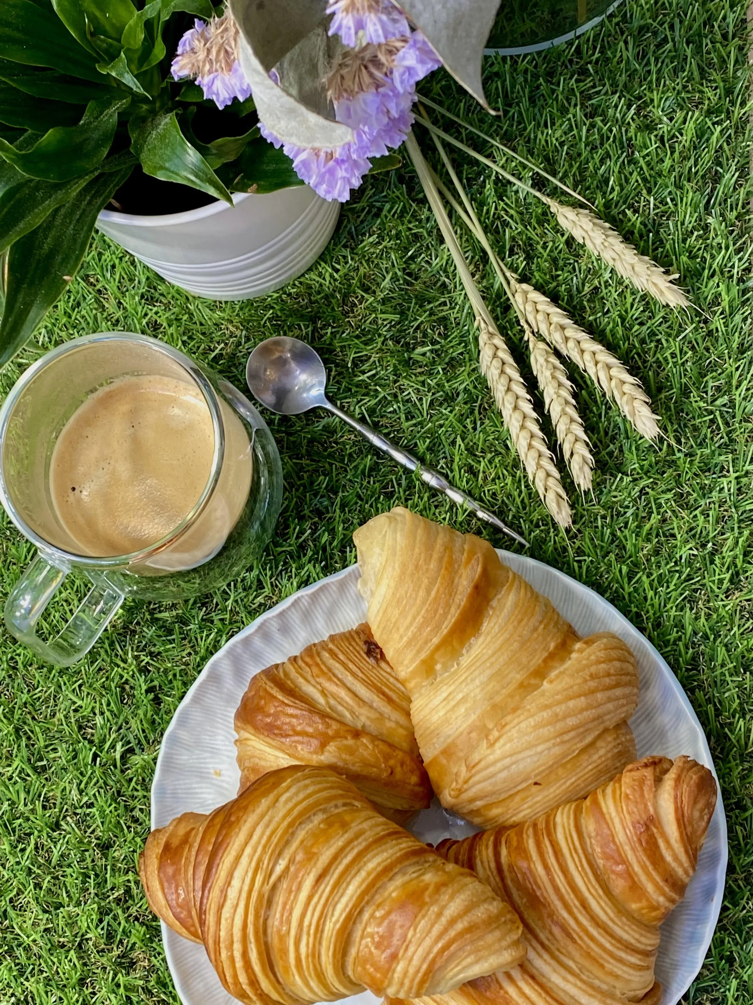 Croissants sur une assiette, tasse de café, bouquet de fleurs violettes dans un vase, herbes et grains de blé sur l'herbe verte.