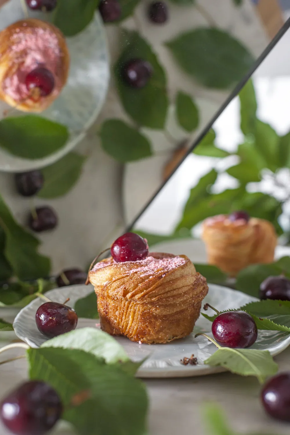 Un muffin à la cerise sur une assiette blanche, entouré de cerises fraîches et de feuilles vertes, avec un miroir derrière reflétant la scène.