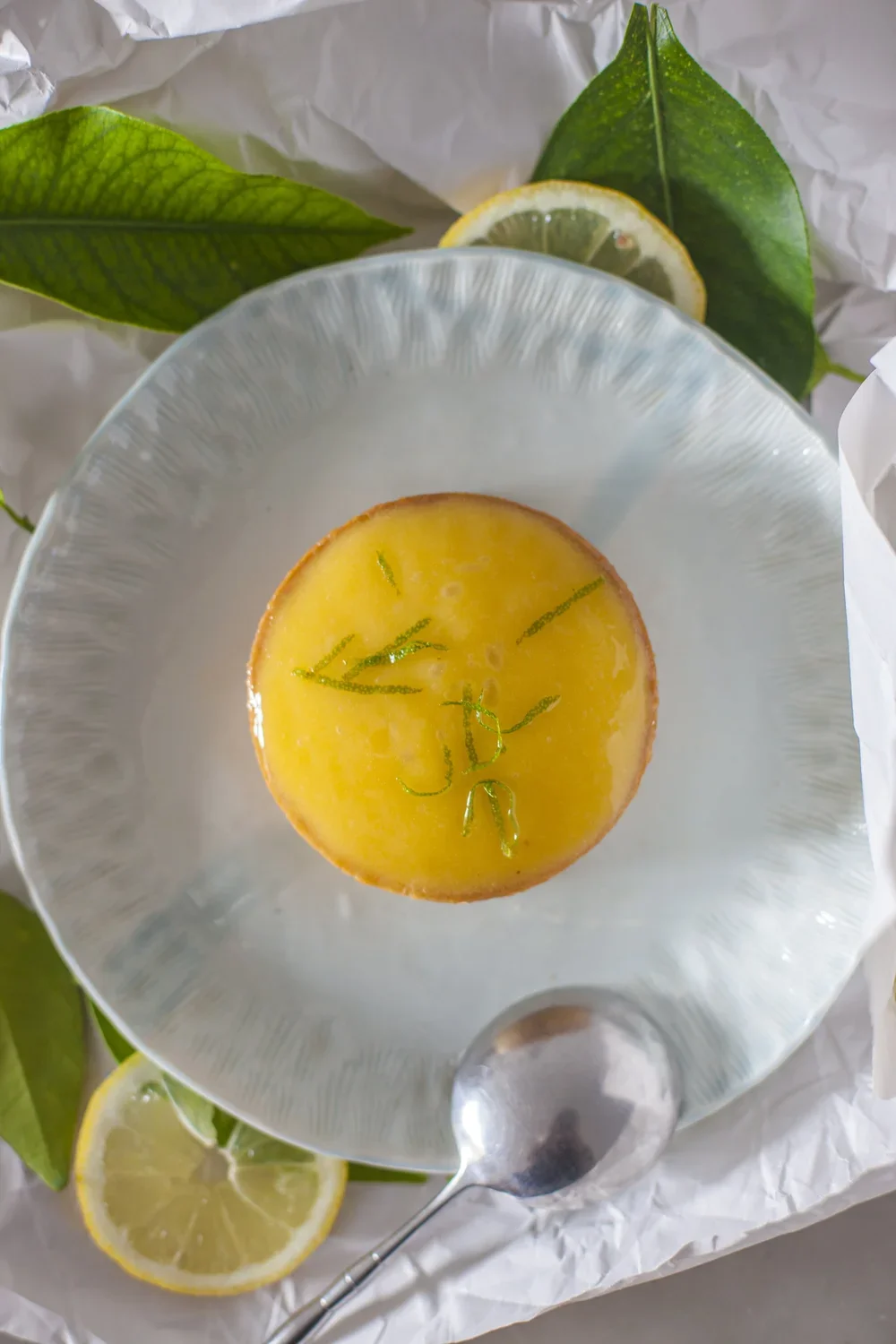 Un gâteau au citron décoré avec des zestes verts, placé dans une coupe en verre, entouré de feuilles vertes et de tranches de citron.