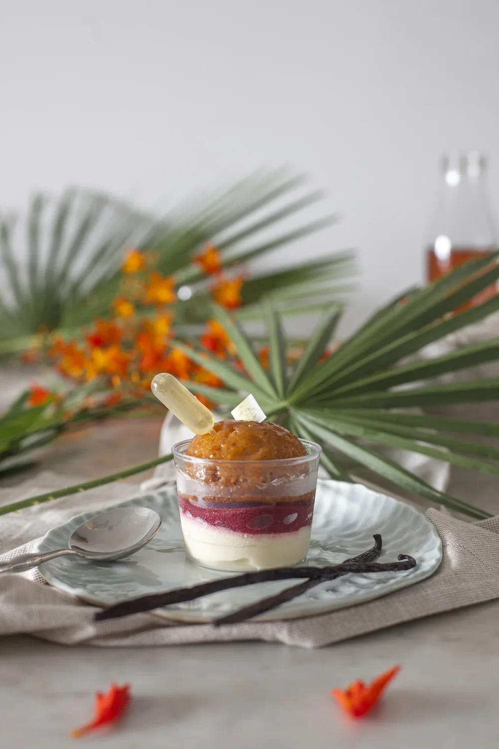 Dessert dans un verre avec mousse caramel, gelée de fruits rouges, crème blanche, décoré d'une gousse de vanille, un biscuit et une cuillère, avec des feuilles de palmier et des fleurs orange en arrière-plan.
