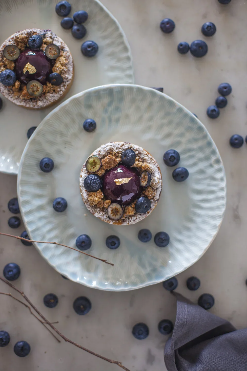 Deux tartelettes aux myrtilles décorées de myrtilles fraîches, de tranches de groseilles, de poudre de sucre et de feuilles d'or, servies sur des assiettes en céramique, entourées de myrtilles dispersées sur une surface claire.