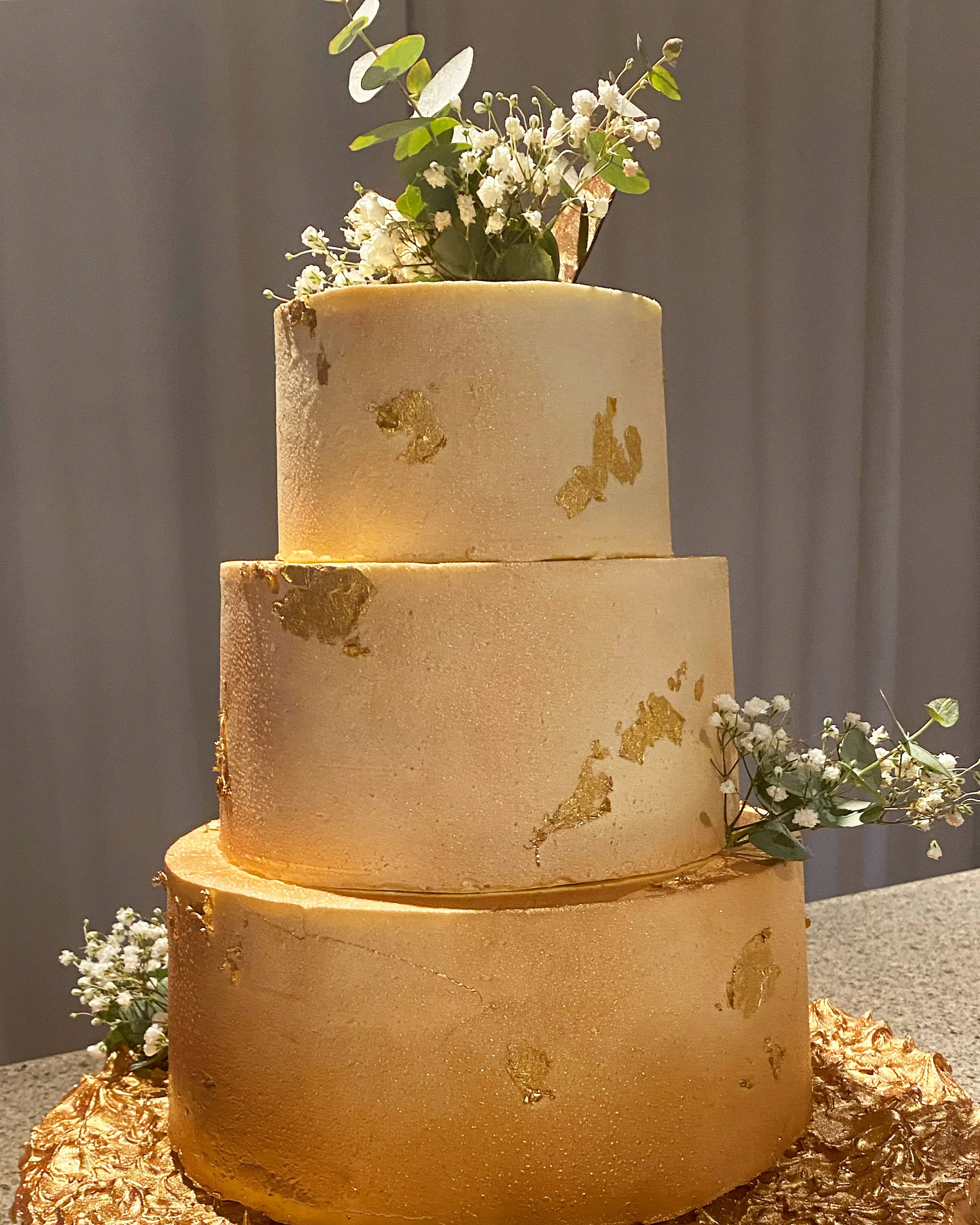 Gâteau à trois étages de couleur dorée avec des décorations en feuilles d'or et des fleurs blanches en haut, posé sur une surface en pierre.