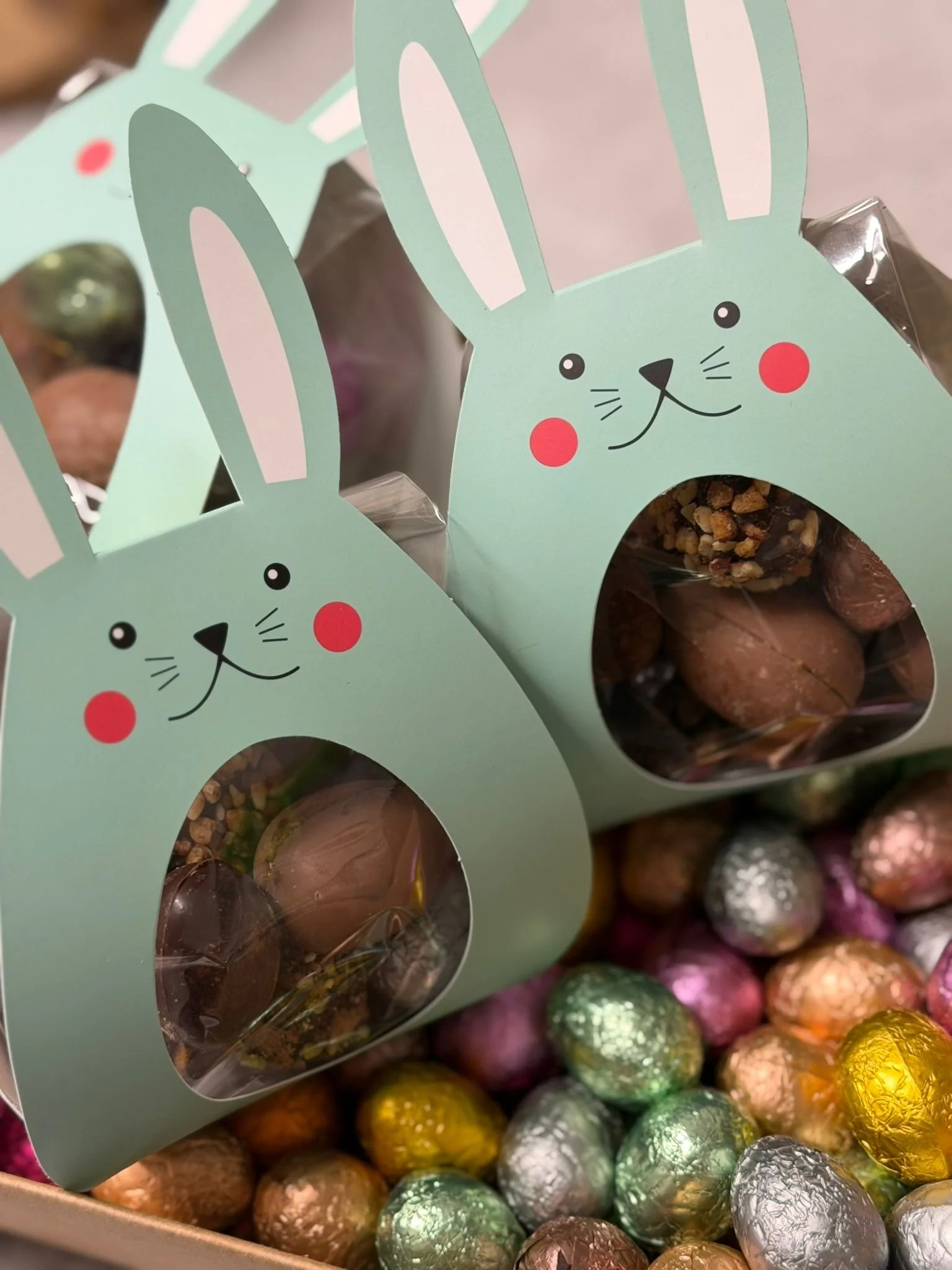 Deux paquets de chocolat en forme de lapins avec un visage de lapin dessinée, entourés de petits œufs en chocolat colorés, dans une boîte.