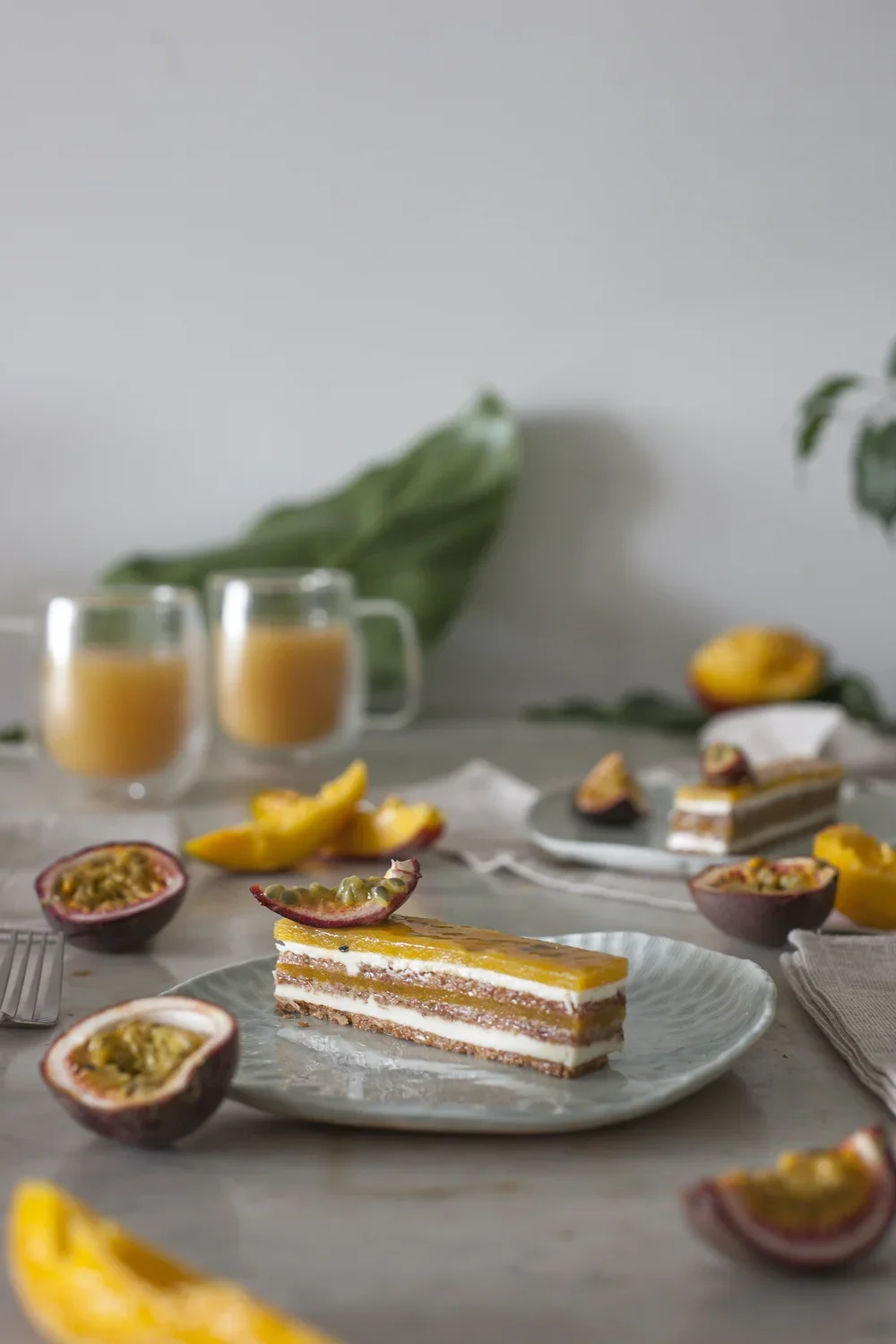 Gâteau aux couches de crème, passion et mangue sur une assiette, avec passion coupée et deux mugs de jus d'orange en arrière-plan, décoration végétale.