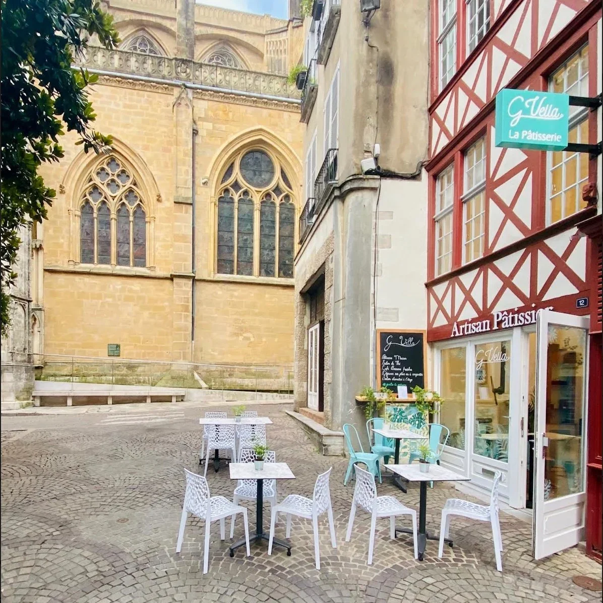 Une petite place pavée avec des tables et chaises blanches devant une pâtisserie appelée G Vella à Strasbourg, avec une église en arrière-plan.