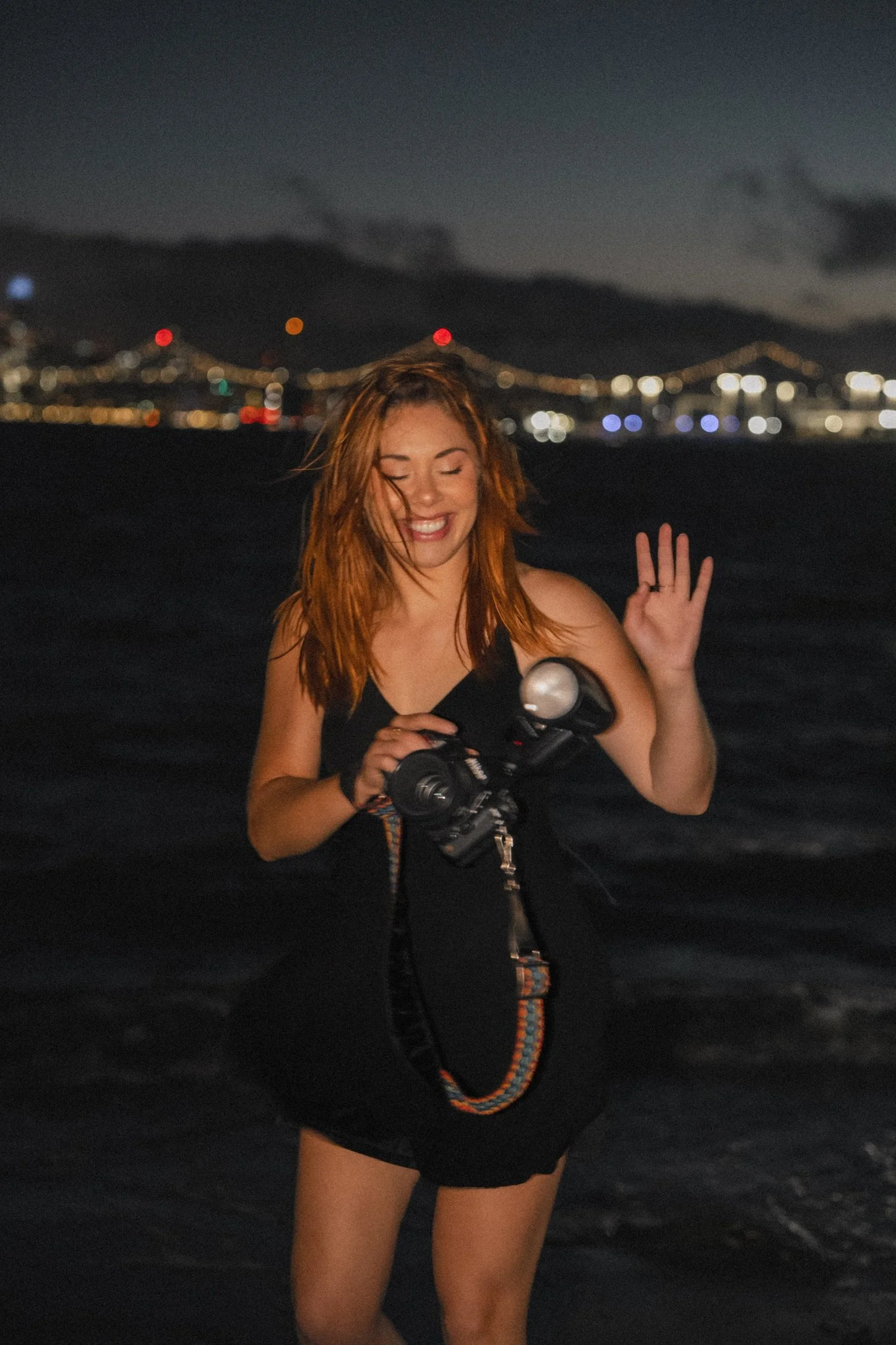 A woman with red hair smiling while holding a camera on a beach at night, with a city and lit-up bridge in the background.