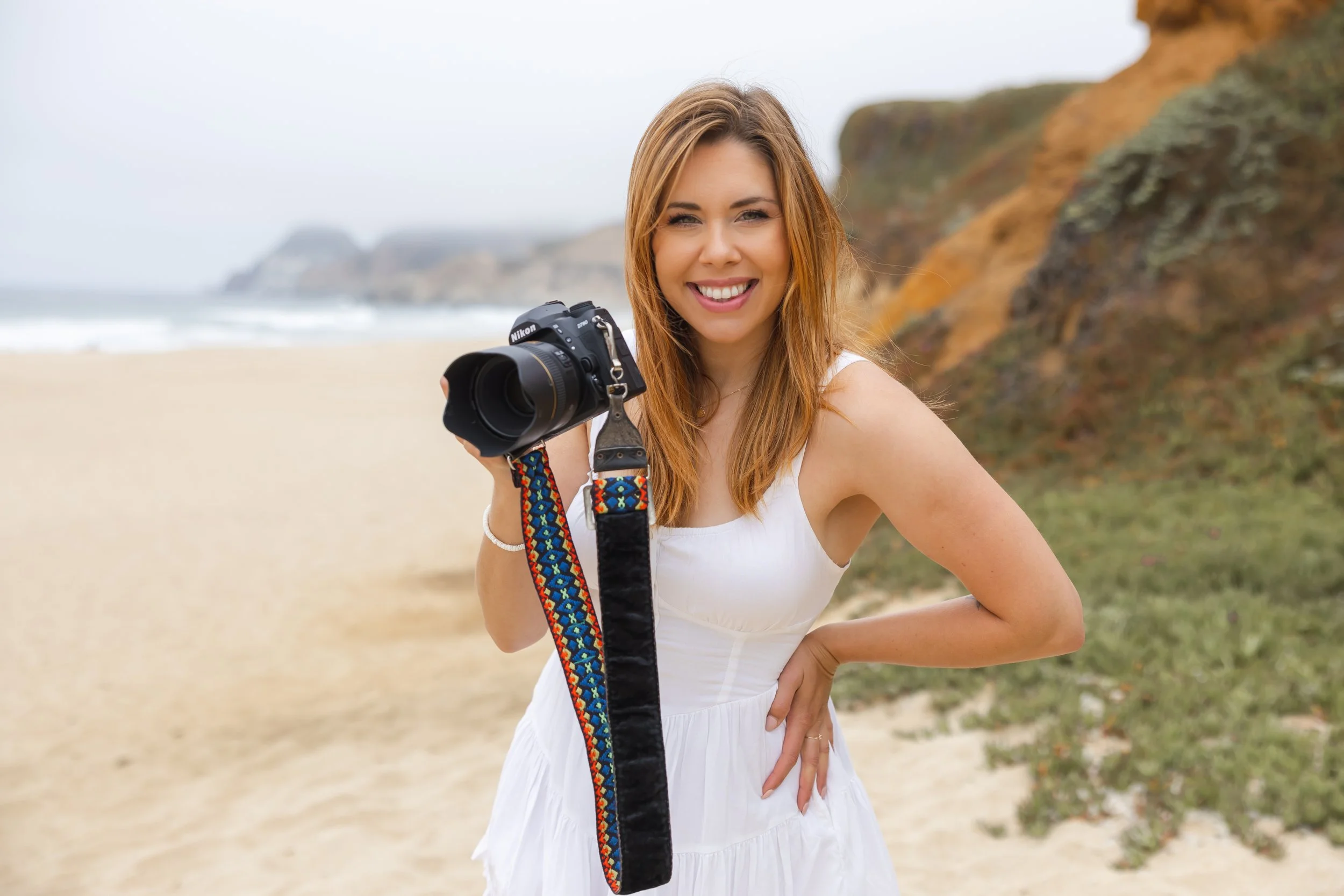 A woman standing on a beach holding a camera, smiling, with sand dunes and the San Diego ocean in the background.