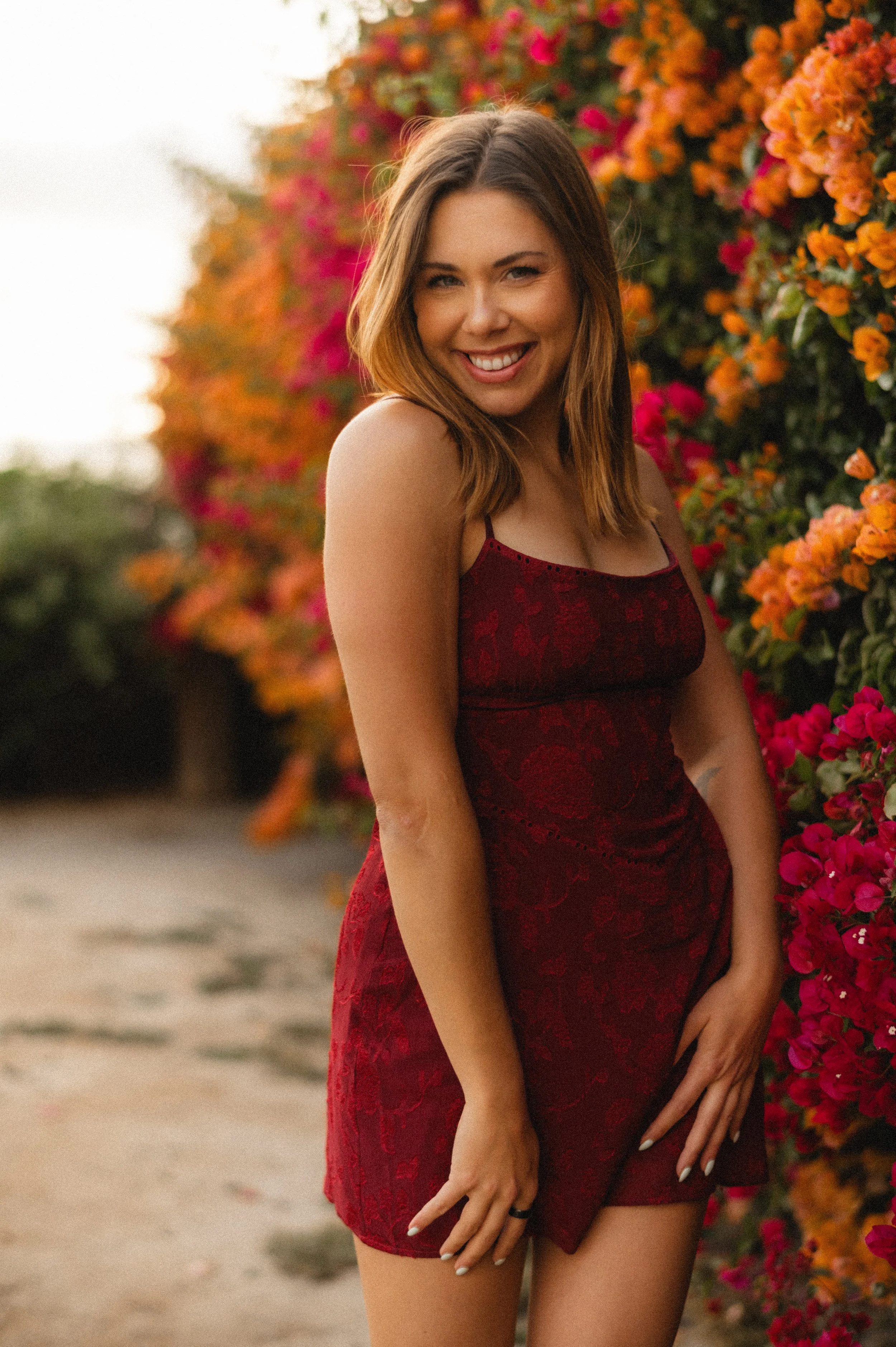 A smiling young woman with shoulder-length brown hair wearing a red lace dress, standing near pink and orange bougainvillea flowers on a sunny day.