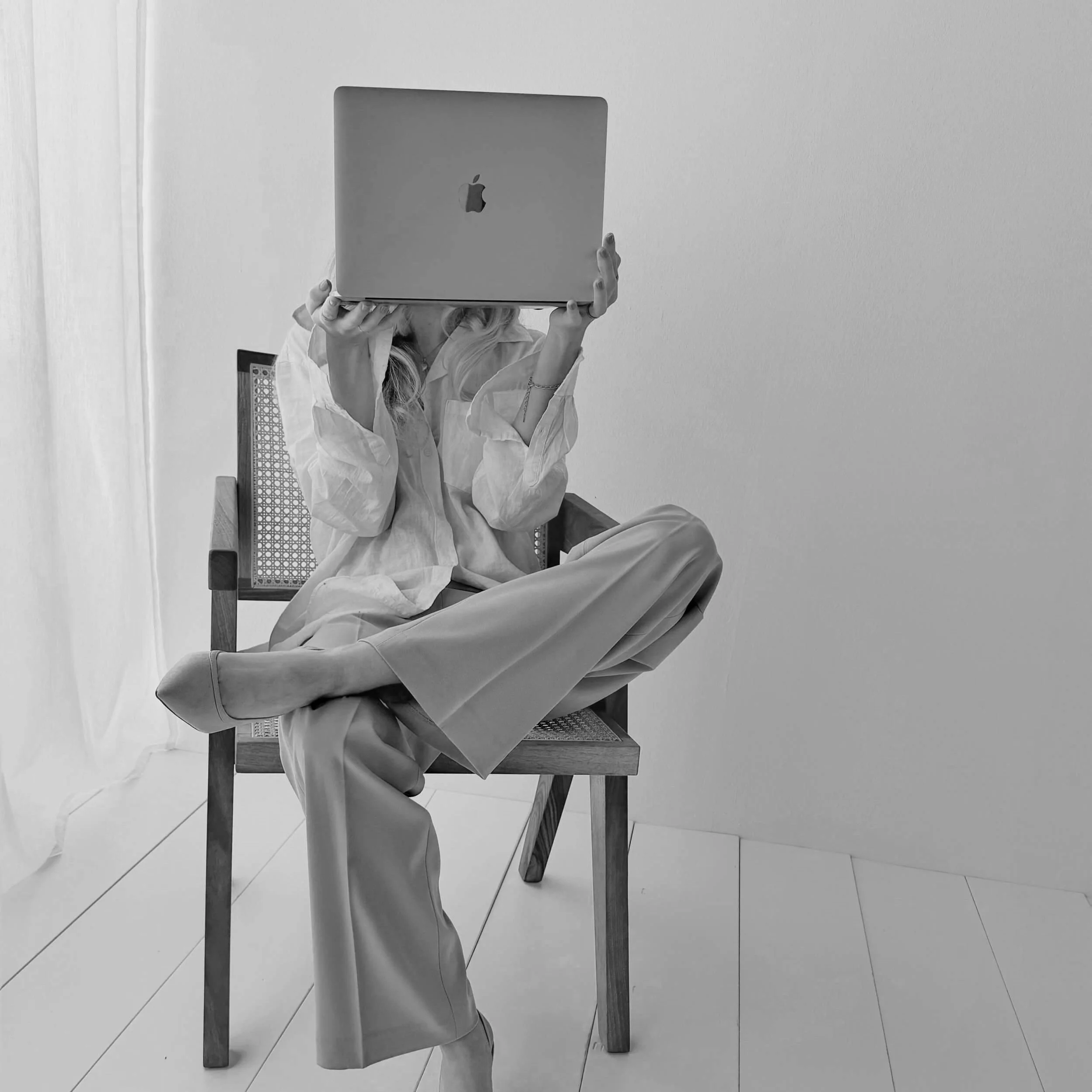 Person sitting cross-legged on a wooden chair, holding a closed MacBook laptop in front of their face, in a minimalist room with white walls and floor.
