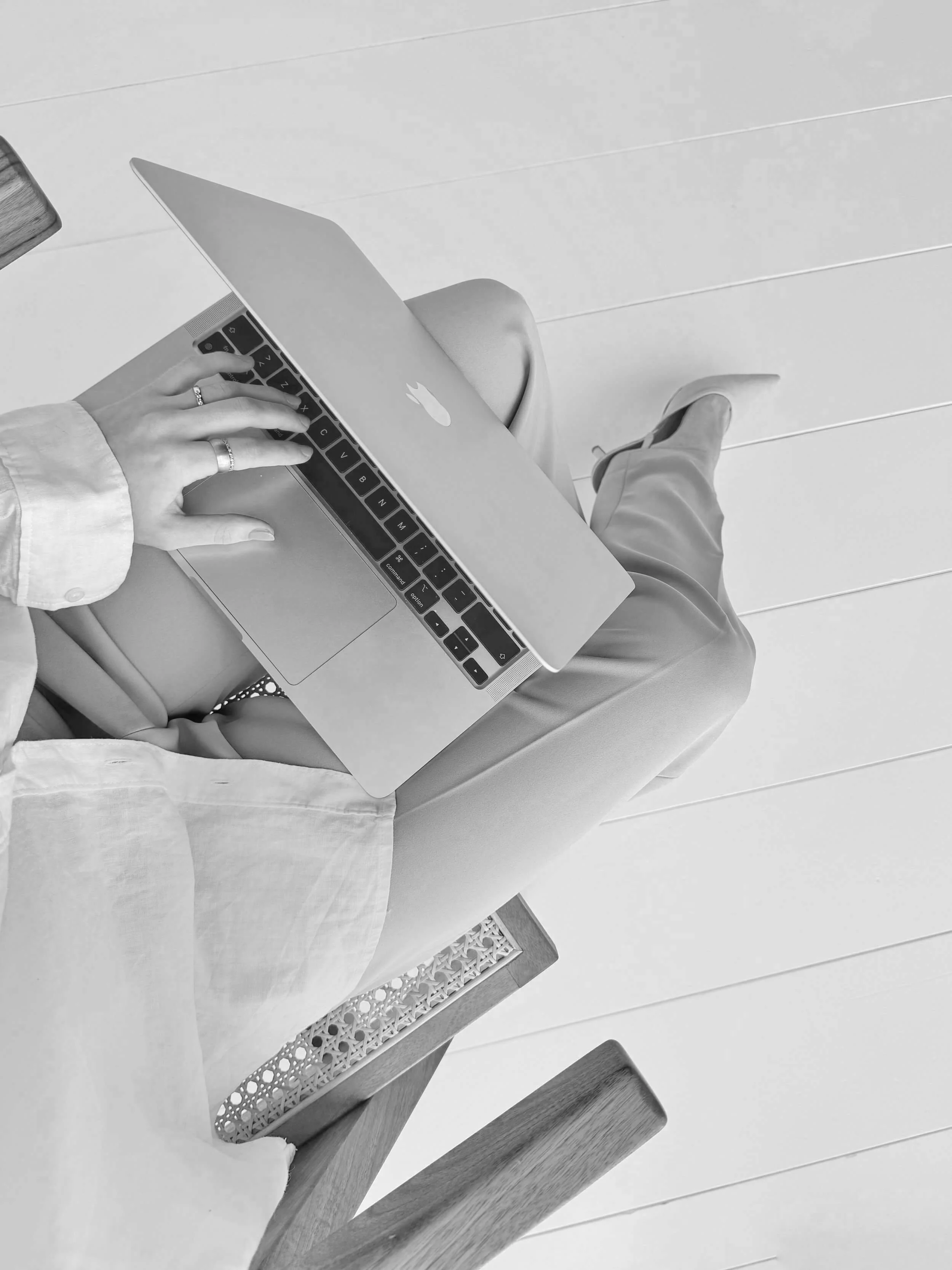 A person using a MacBook with a visible apple logo, sitting on a chair with a woven backrest, on a wooden floor.