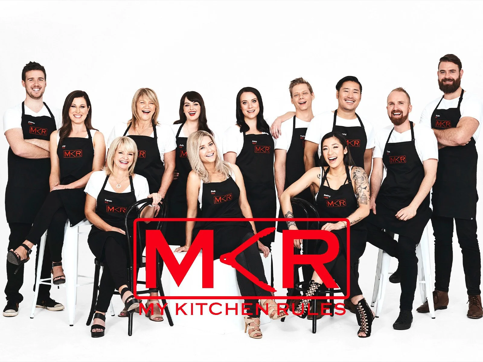 Group of fifteen people in black aprons with red MKR logo, smiling and posing, some sitting and some standing, against a white background.